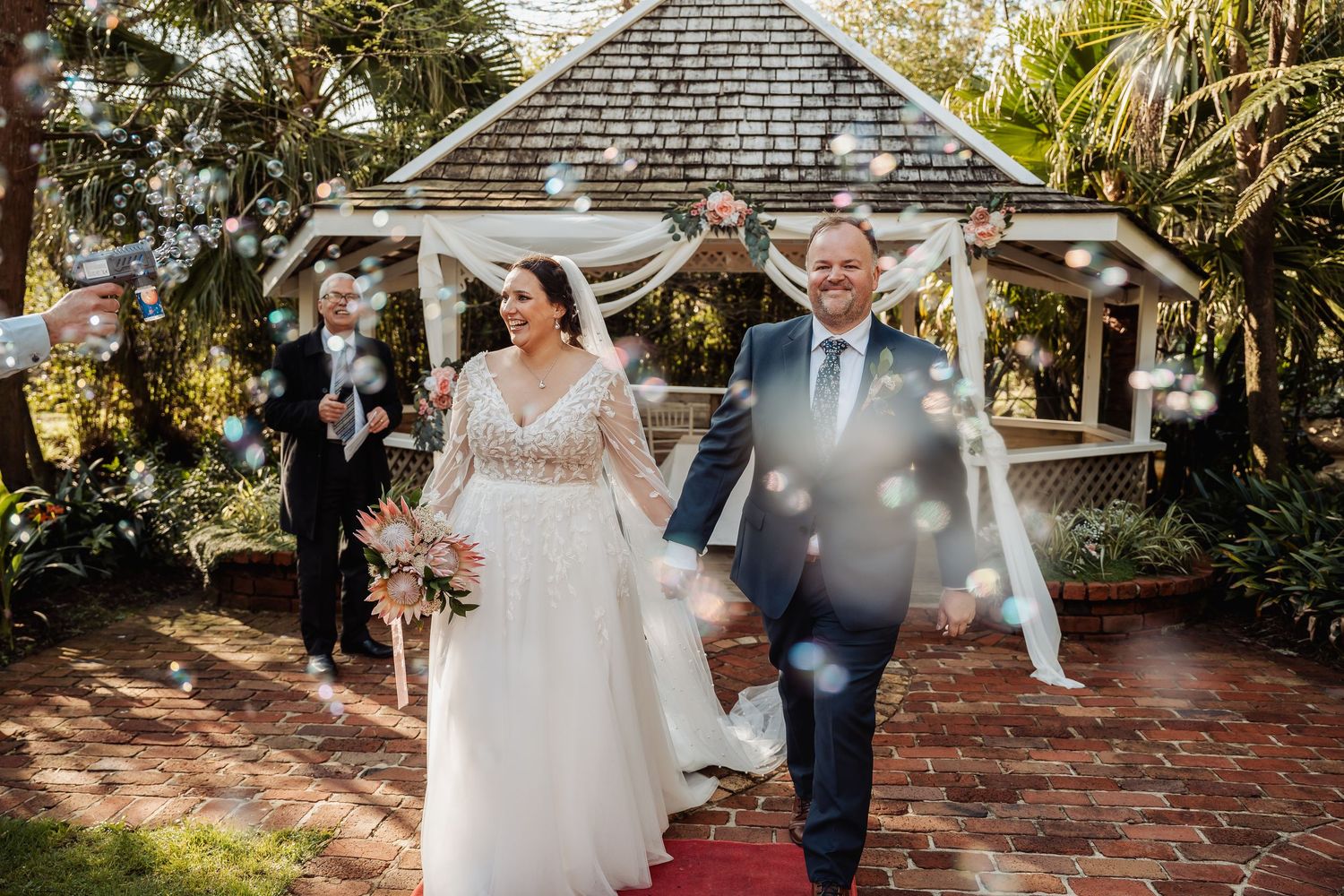 Newlyweds exit their garden wedding ceremony under a shower of confetti near a gazebo.