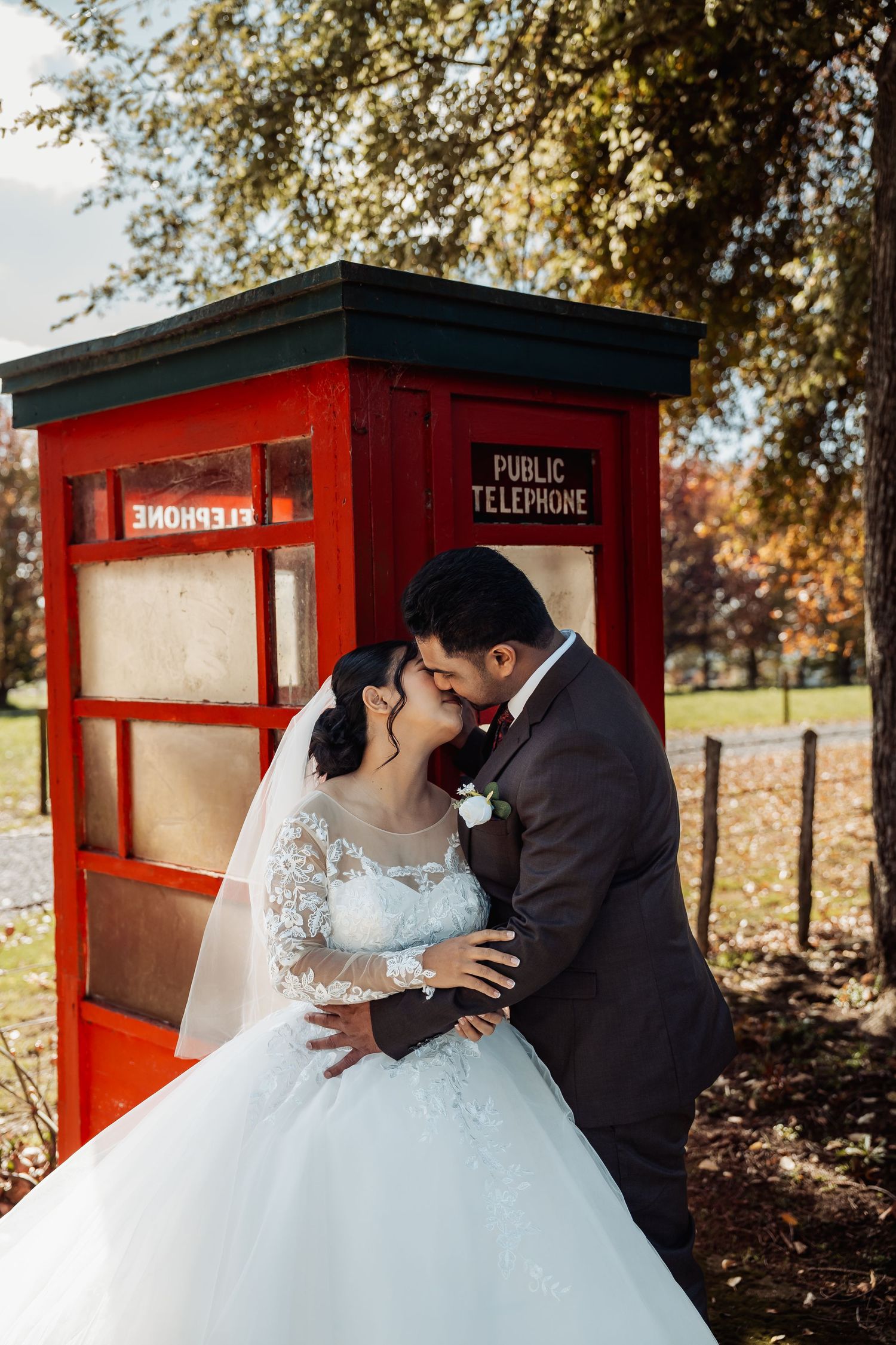 Bride and groom sharing a romantic kiss beside a red public telephone booth in autumn park.