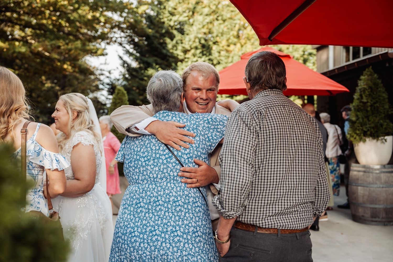 Wedding guests embrace and celebrate outdoors near rustic barn venue under red umbrellas.