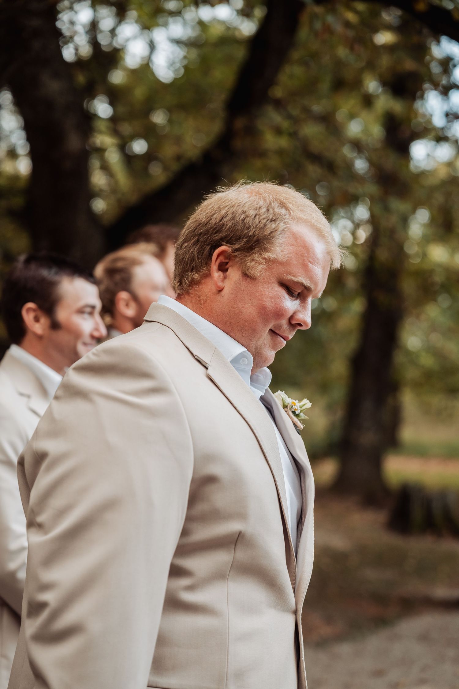 Groom in a tan suit with boutonniere standing outdoors among trees with groomsmen in background.
