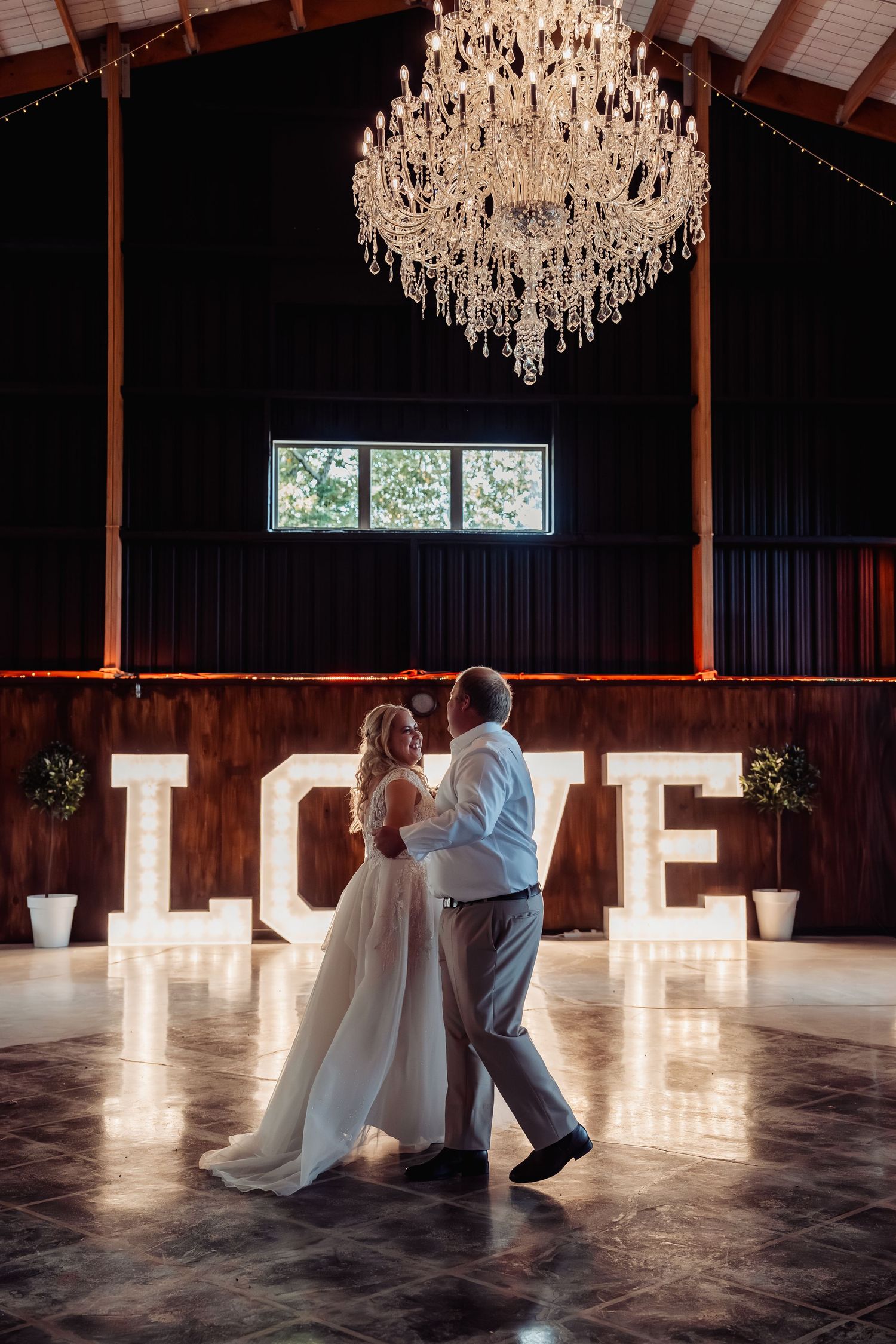 Bride and groom share first dance under crystal chandelier in front of illuminated LOVE letters in rustic barn venue.