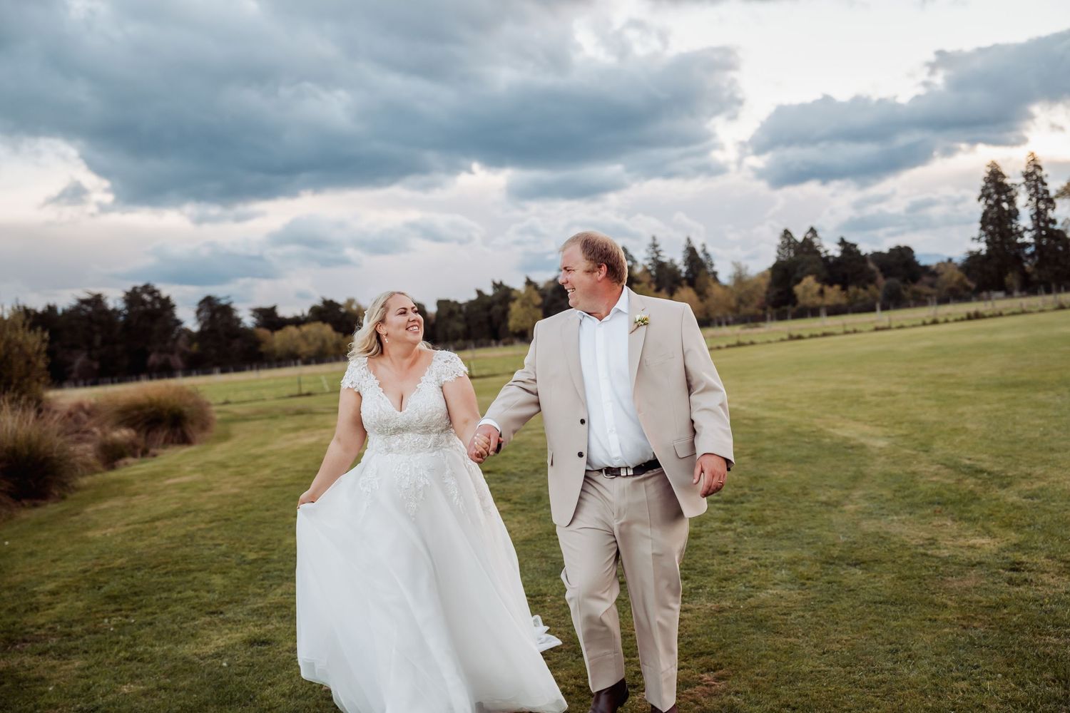 Bride and groom walk hand in hand across green field under dramatic cloudy sky.