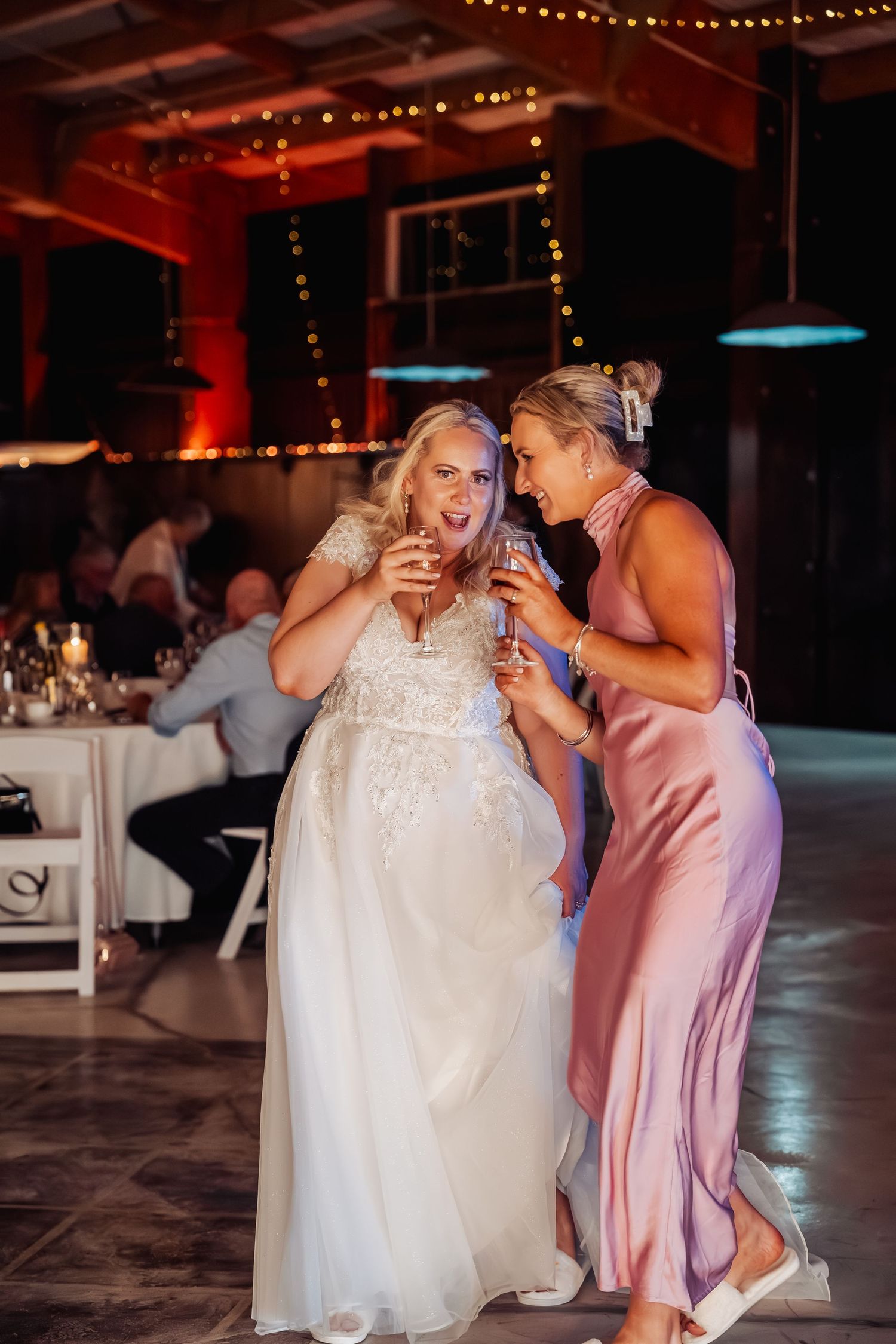 Bride in white lace gown toasting drinks with bridesmaid in pink dress at wedding reception.