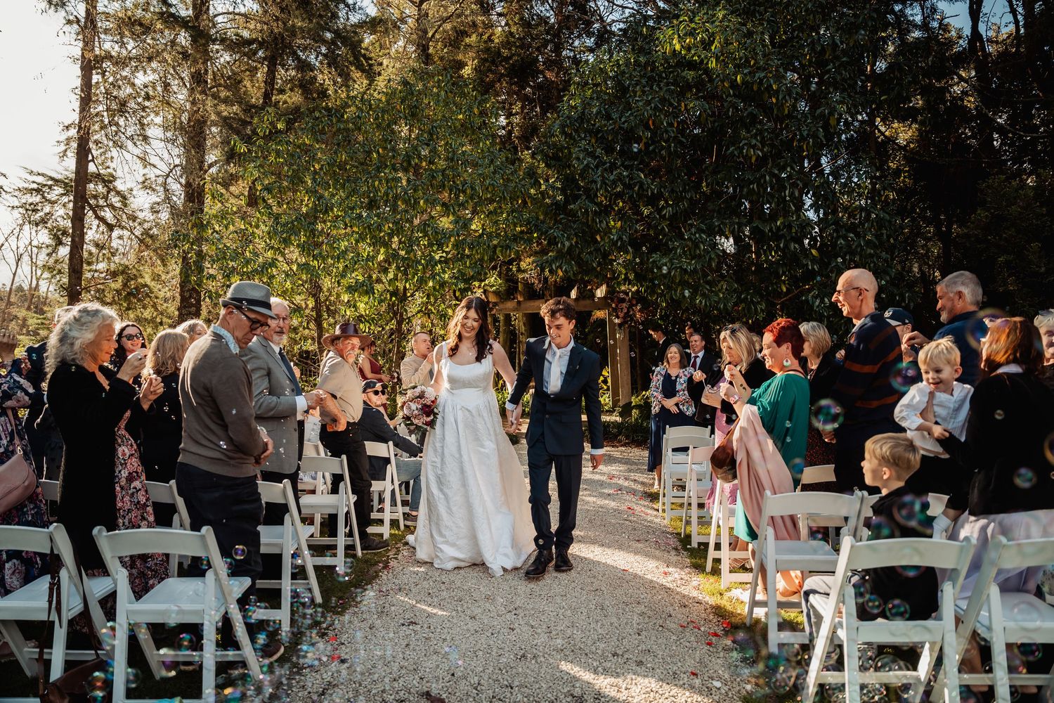 Bride and groom walking down gravel aisle surrounded by applauding guests at garden wedding.