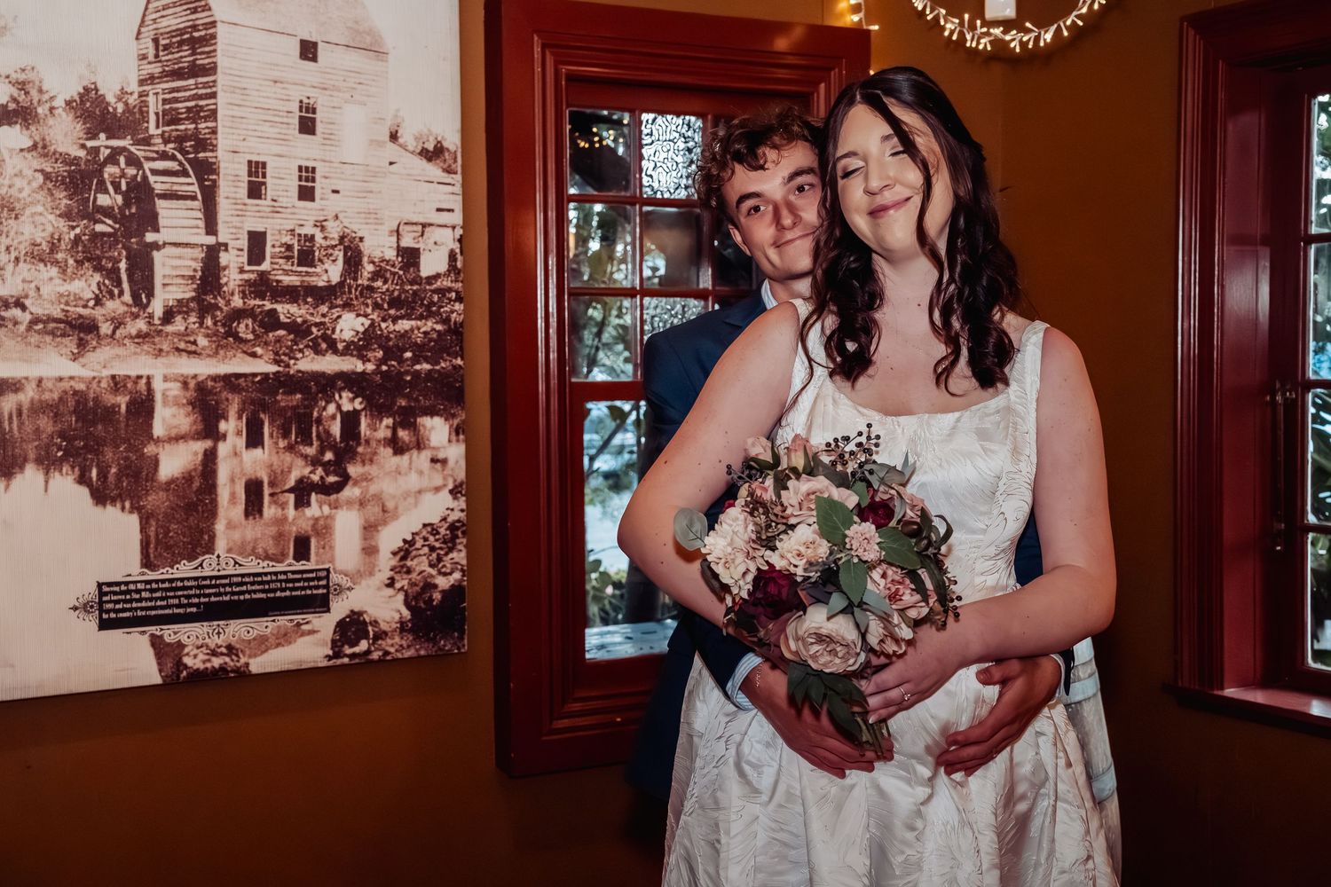 Bride and groom pose together holding bouquet inside rustic venue with vintage wall decor.
