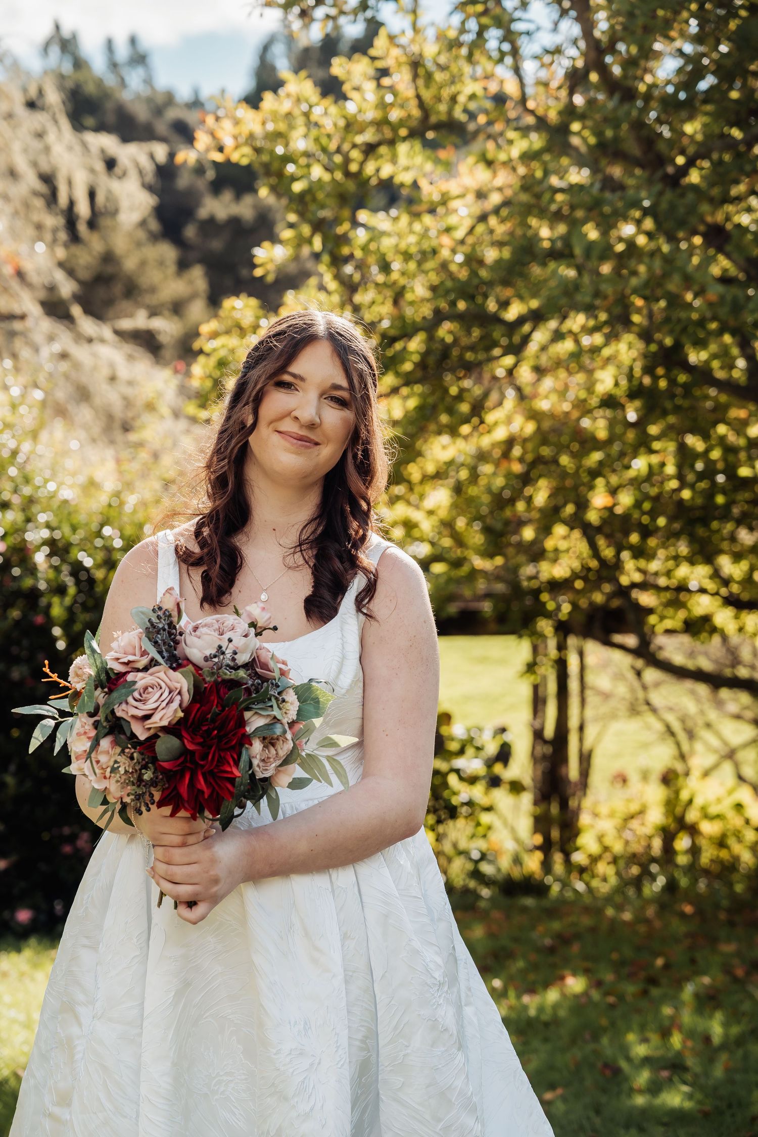 Bride in white gown holding bouquet of pink and red roses smiling in sunlit garden.