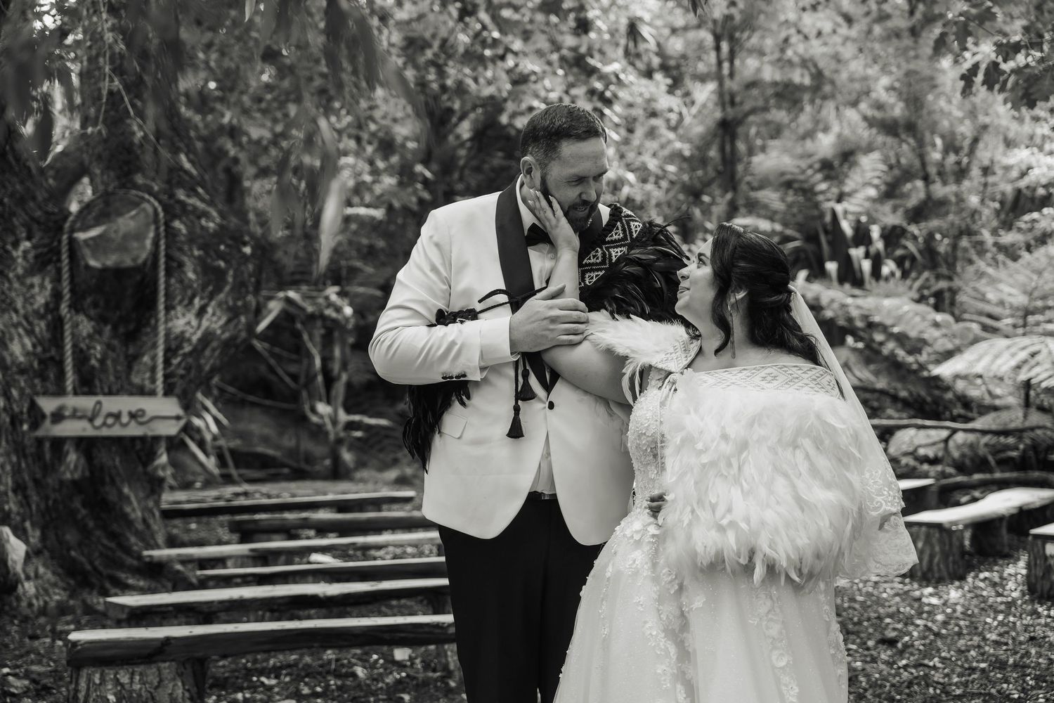 Black and white photo of a bride and groom sharing a tender moment outdoors, surrounded by lush greenery.