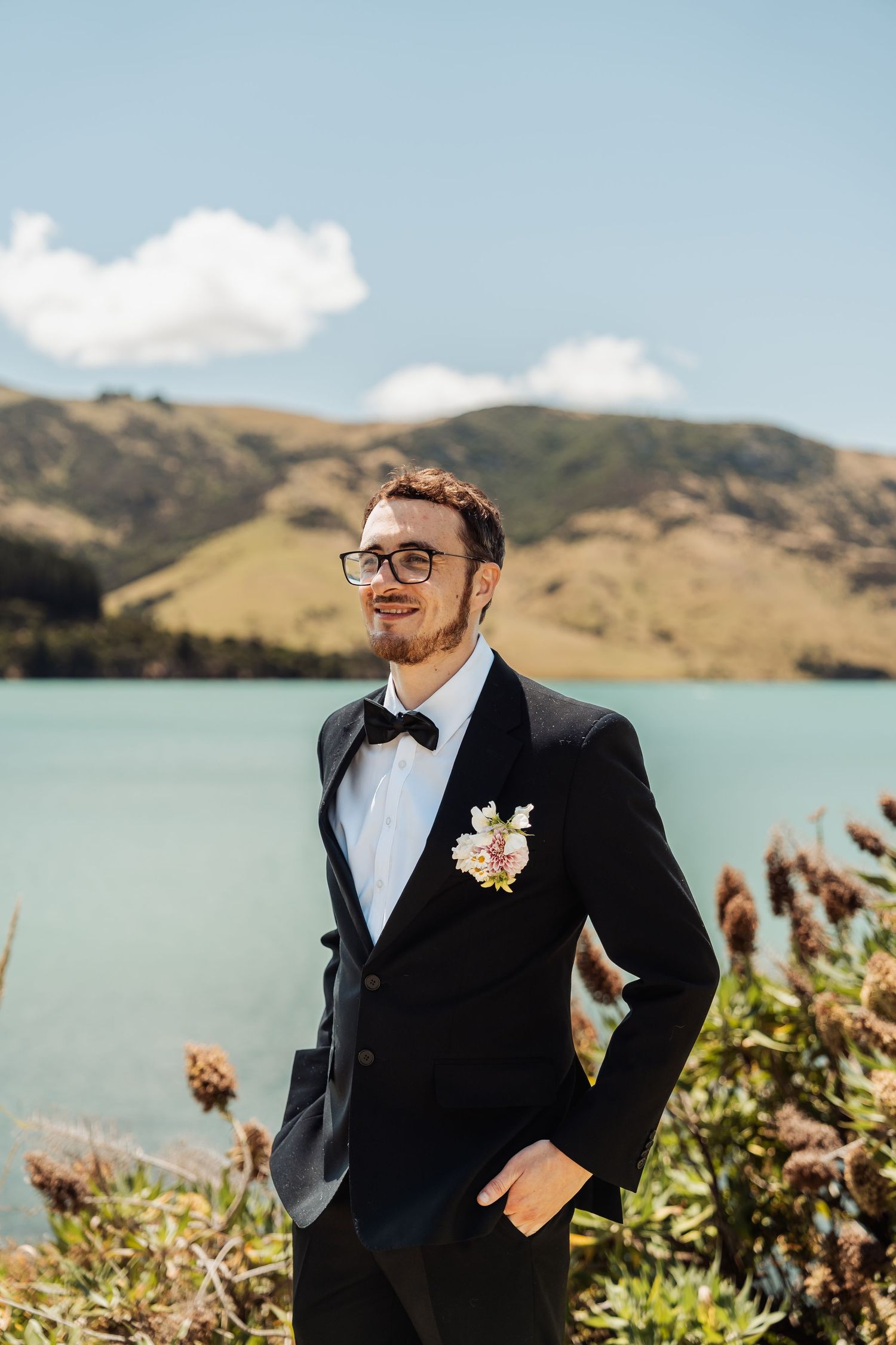 Handsome groom in black tuxedo with boutonniere and bow tie standing by turquoise lake and mountains.