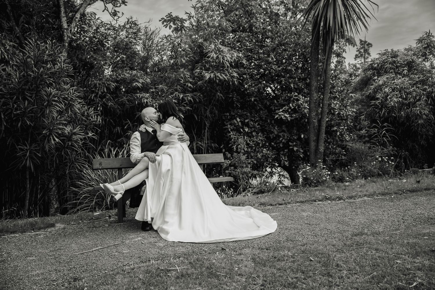 Black and white photo of bride sitting on groom's lap sharing a kiss on a garden bench.
