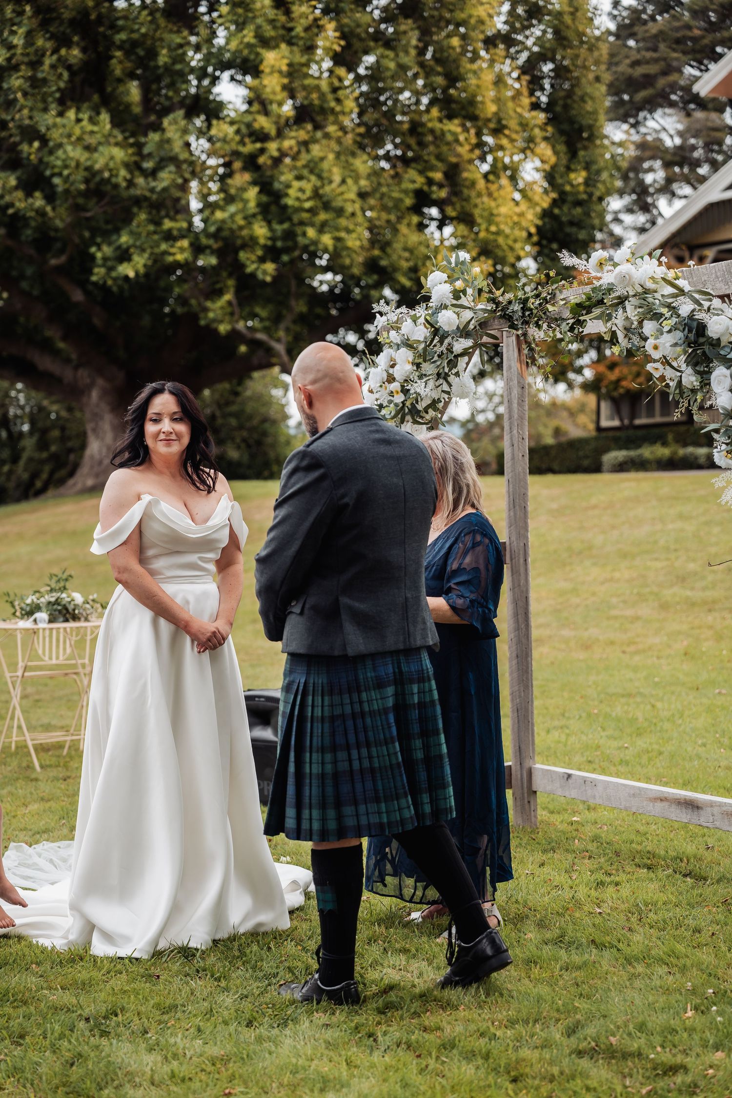 Bride in off-shoulder white gown exchanges vows with groom in kilt at outdoor ceremony.