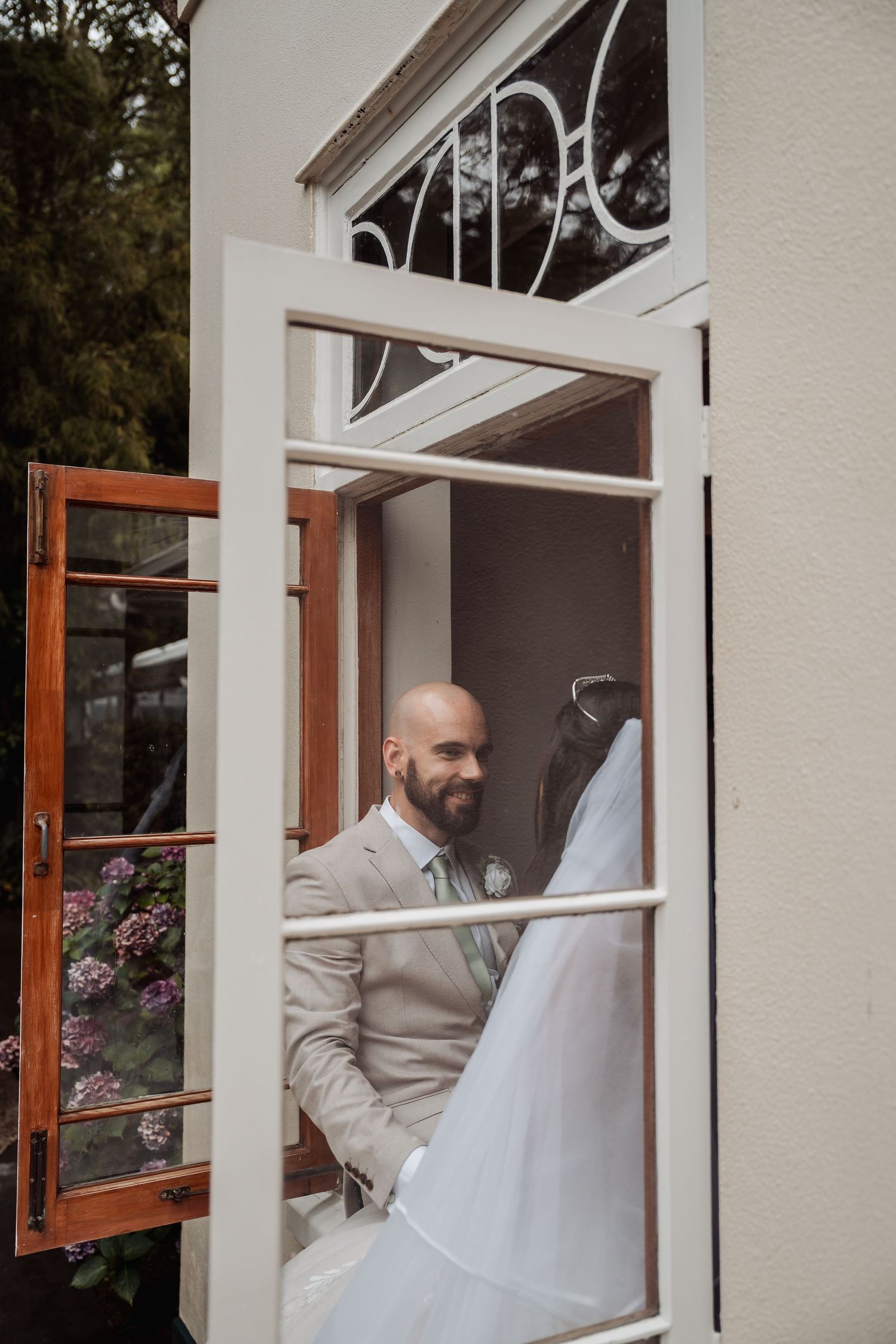Smiling bald groom in beige suit seen through open window with bride's veil in foreground.