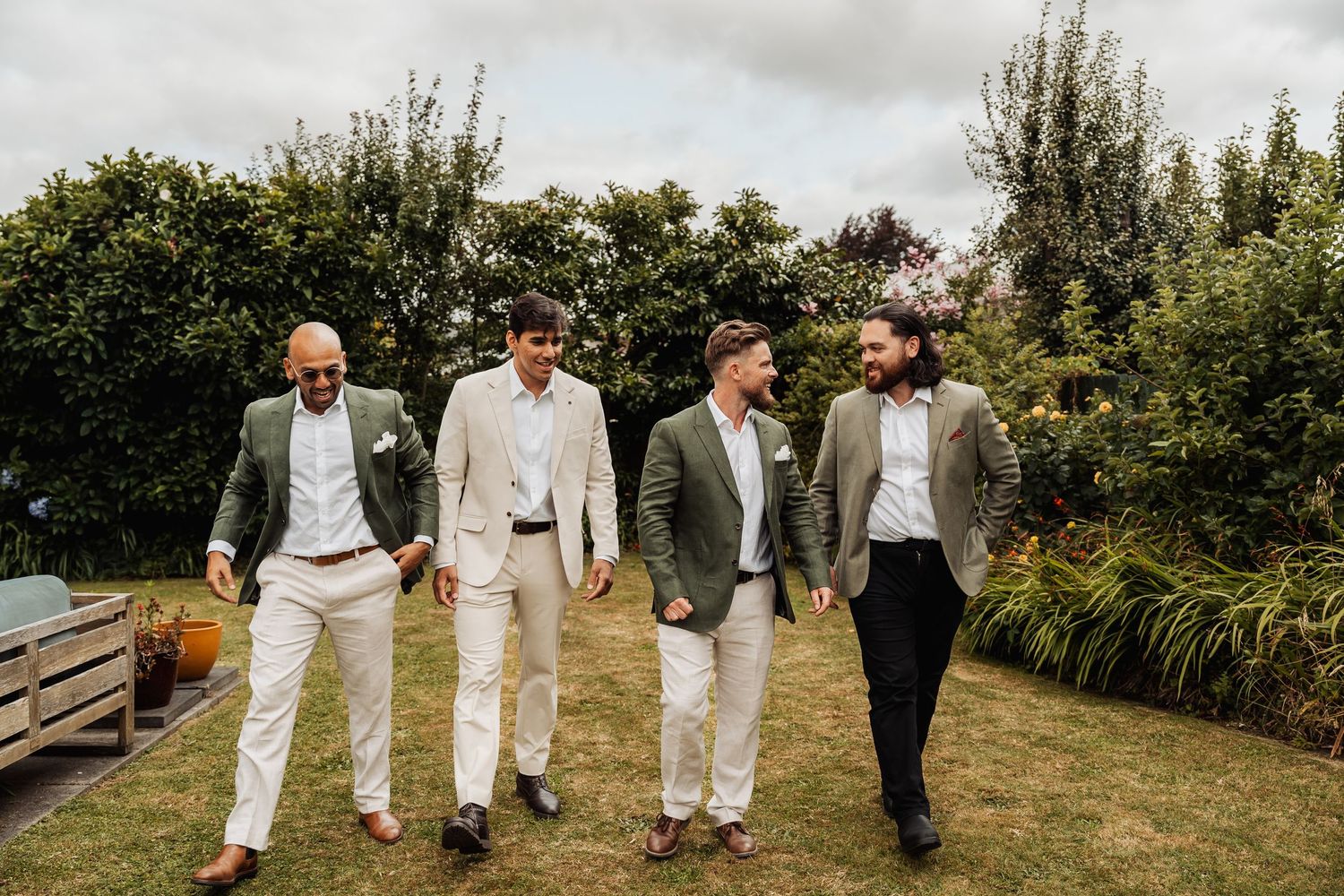 Four groomsmen in suits walking together on a green lawn at an outdoor wedding celebration.