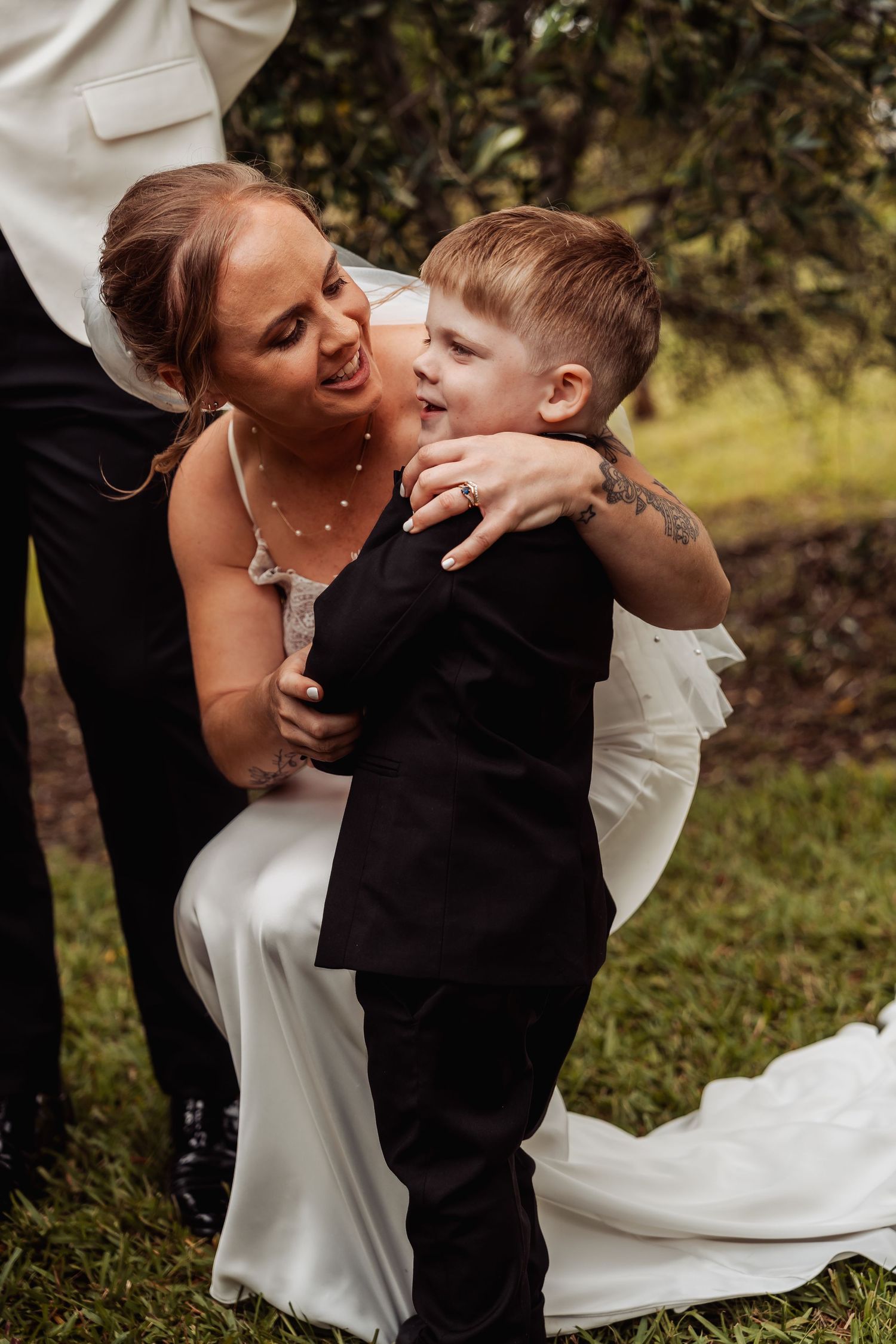 Bride in white gown hugging young boy in black suit during wedding celebration.