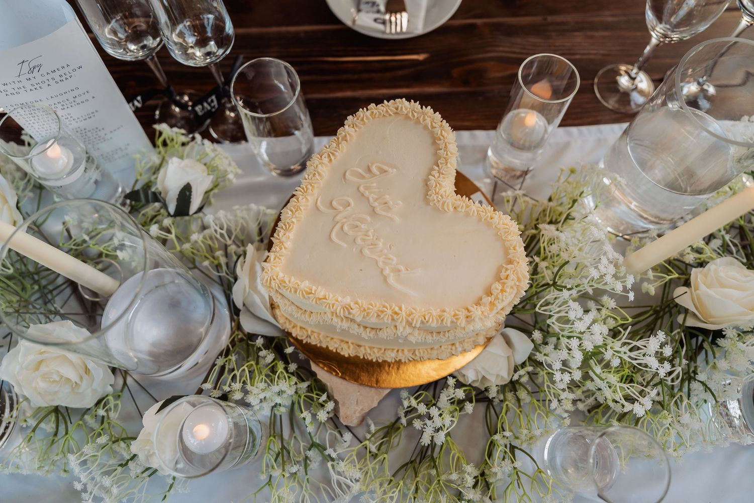 Heart-shaped wedding cake on floral table with white roses, candles, and elegant glassware.