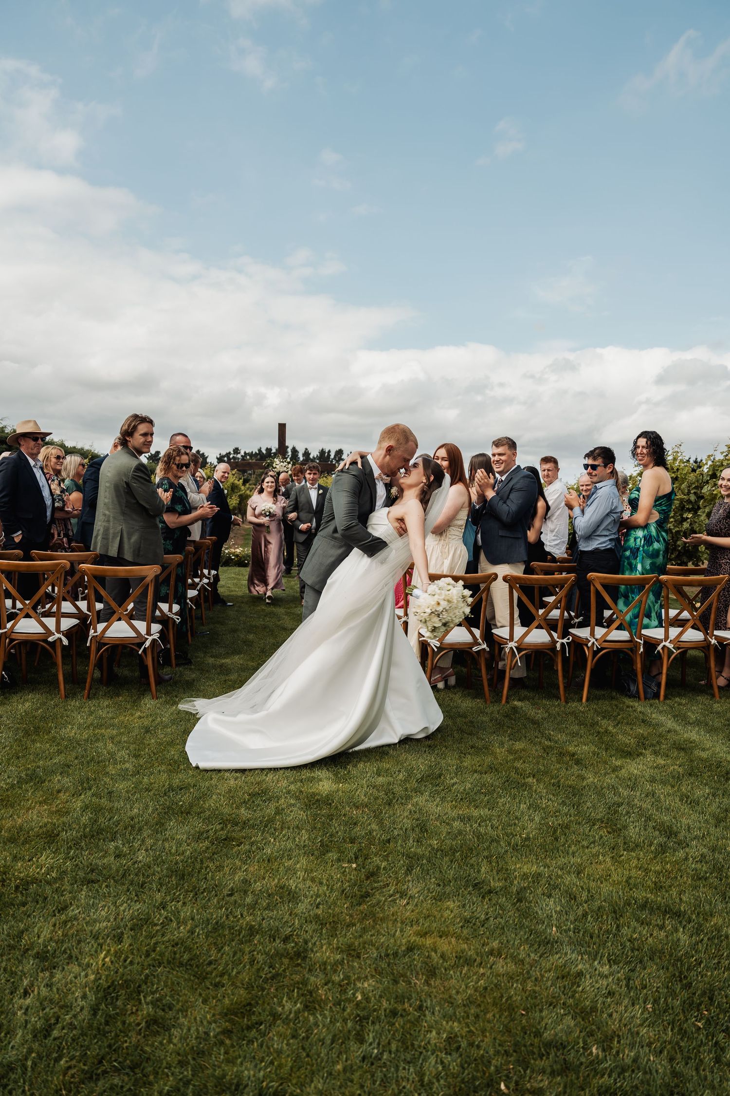 Bride and groom share a romantic dip kiss at outdoor wedding ceremony while guests applaud on a sunny day.