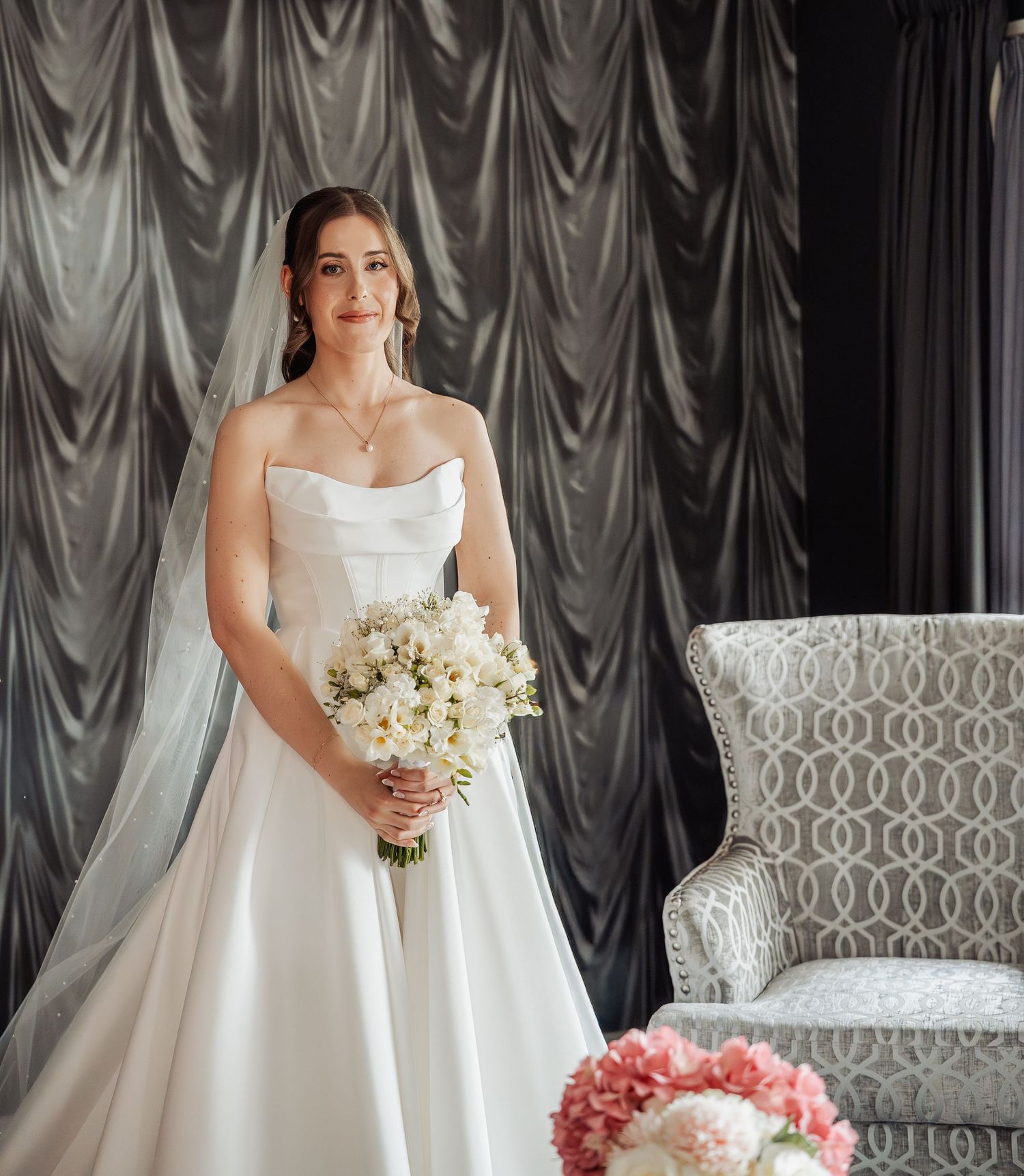 Bride in strapless white ball gown holding white floral bouquet, wearing veil, standing before grey draped backdrop.