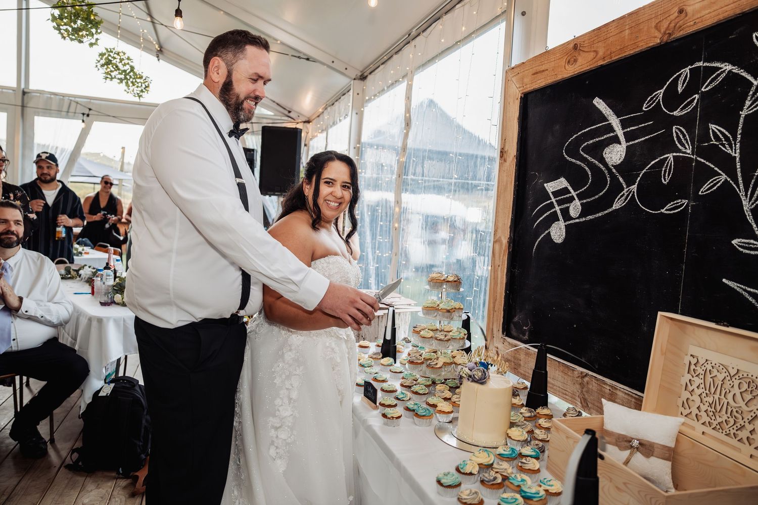 Bride and groom smiling together at wedding cake table during indoor reception celebration.