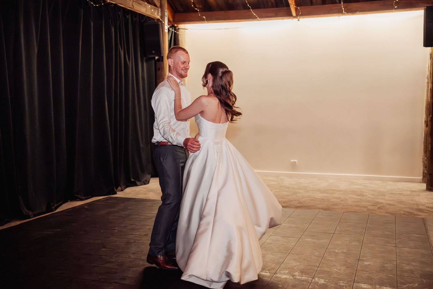 Bride and groom share their first dance in a rustic barn venue, bride in flowing white ballgown.