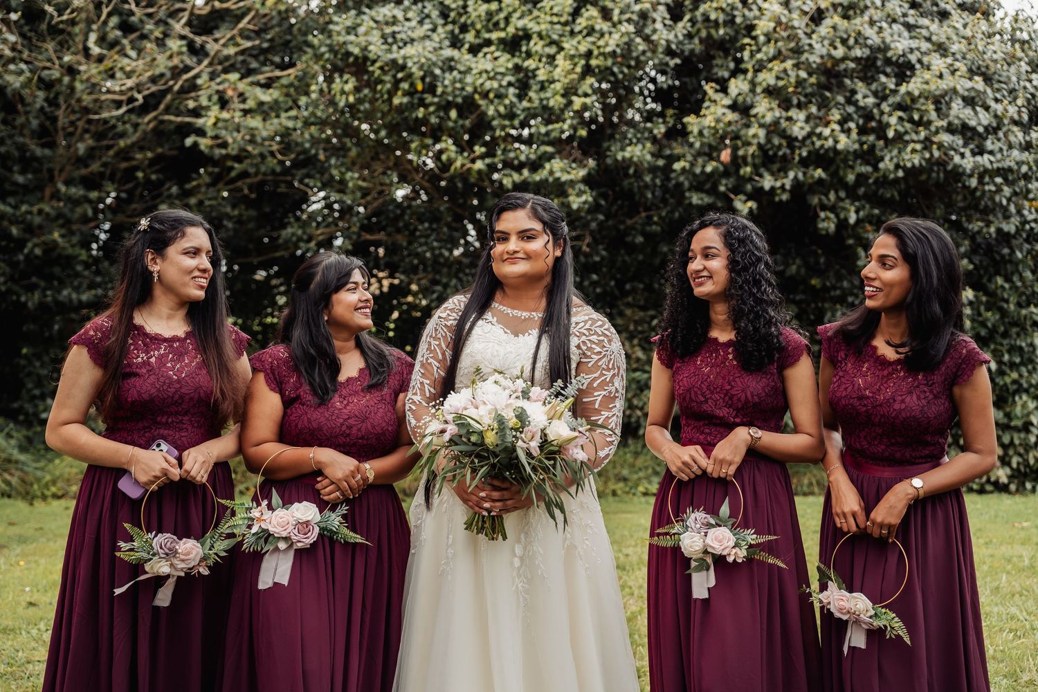 Bride in white gown holds bouquet flanked by four bridesmaids in burgundy dresses outdoors.