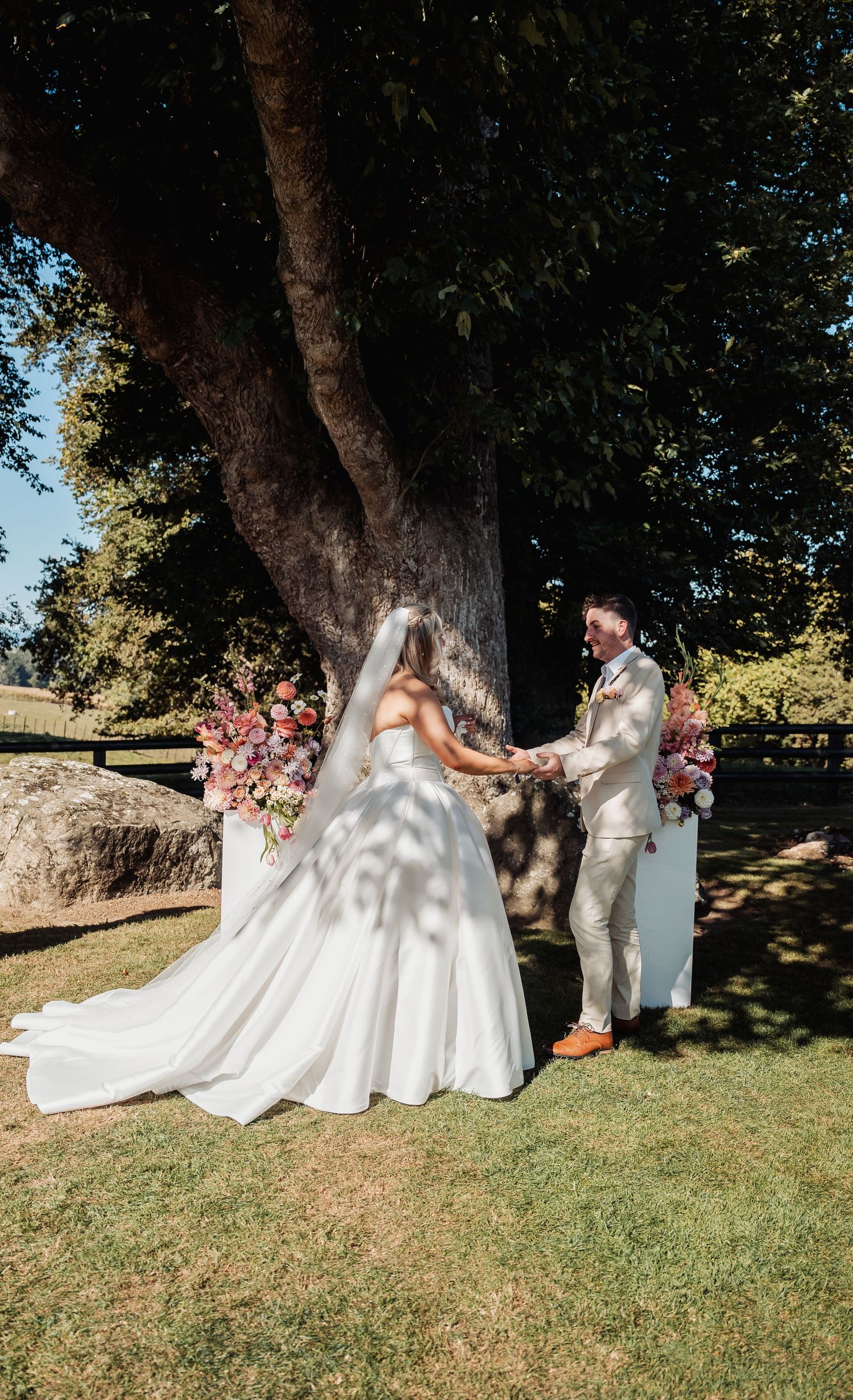 Bride and groom hold hands beneath a large tree during their outdoor wedding ceremony.