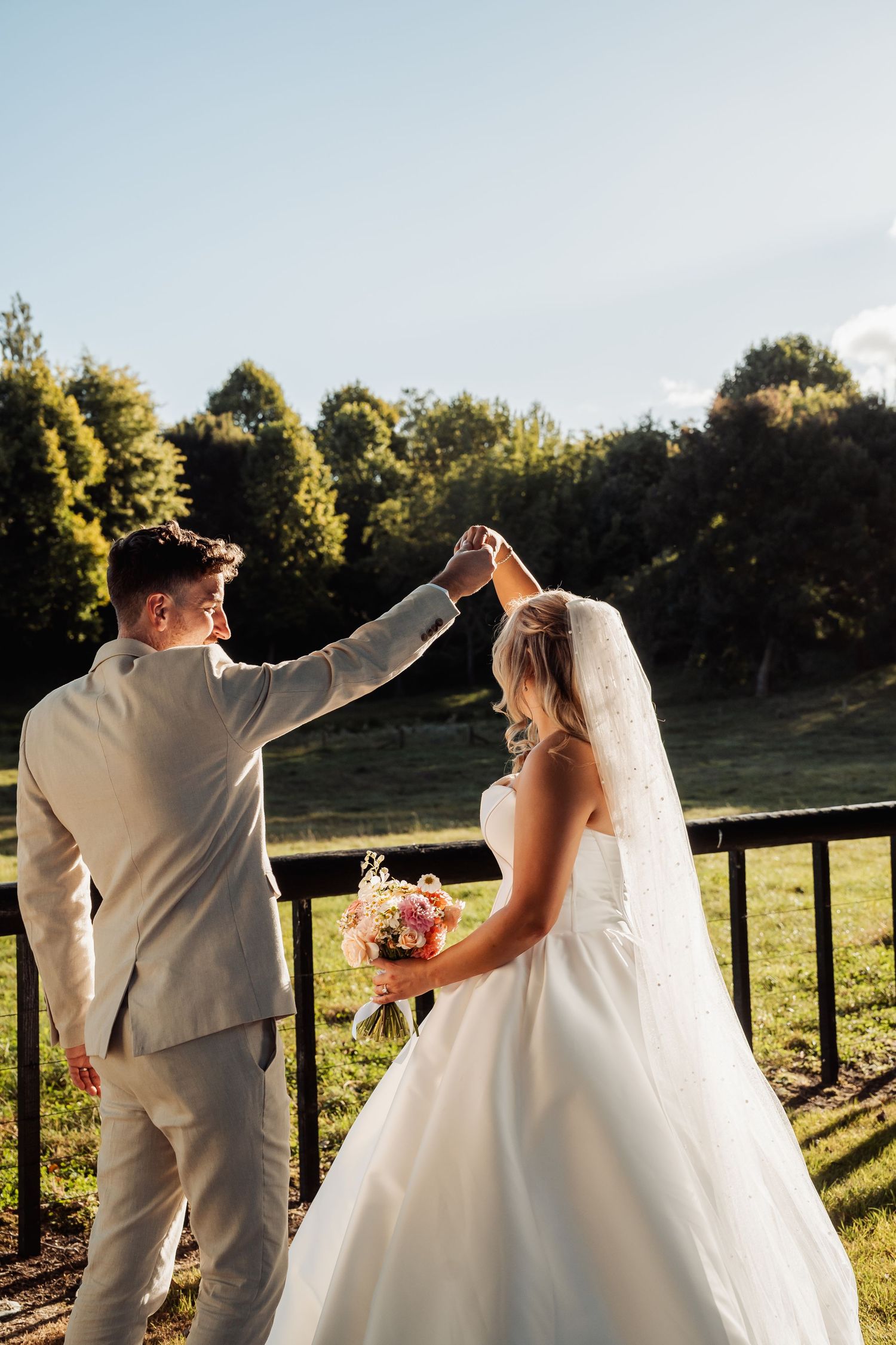 Groom twirls bride in white gown and veil outdoors, holding bouquet, with green trees and blue sky behind.