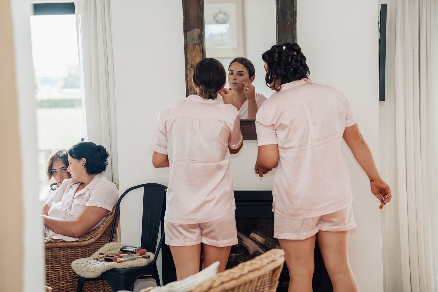 Bridesmaids in pink pajamas helping bride with makeup during wedding morning preparations.