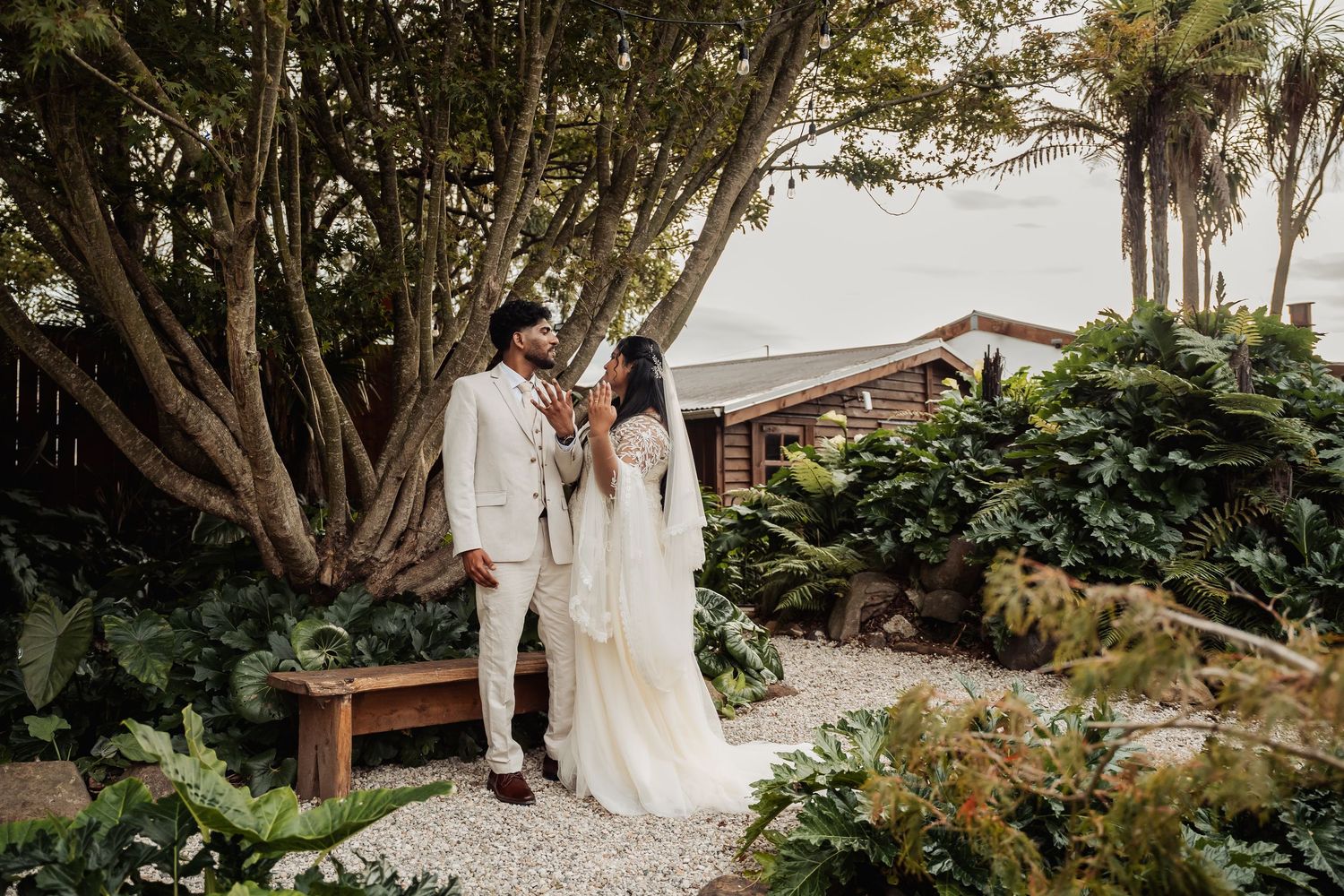 Bride and groom in elegant white attire share a tender moment among tropical garden greenery.