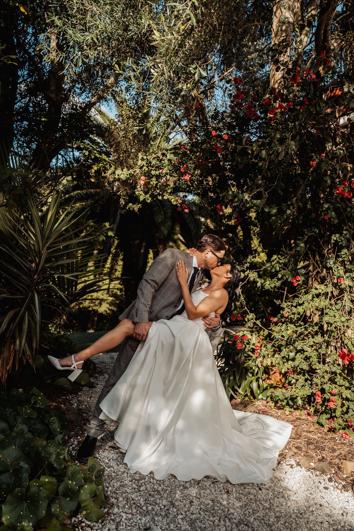 Groom dips bride for romantic kiss surrounded by lush colorful garden greenery after wedding ceremony.
