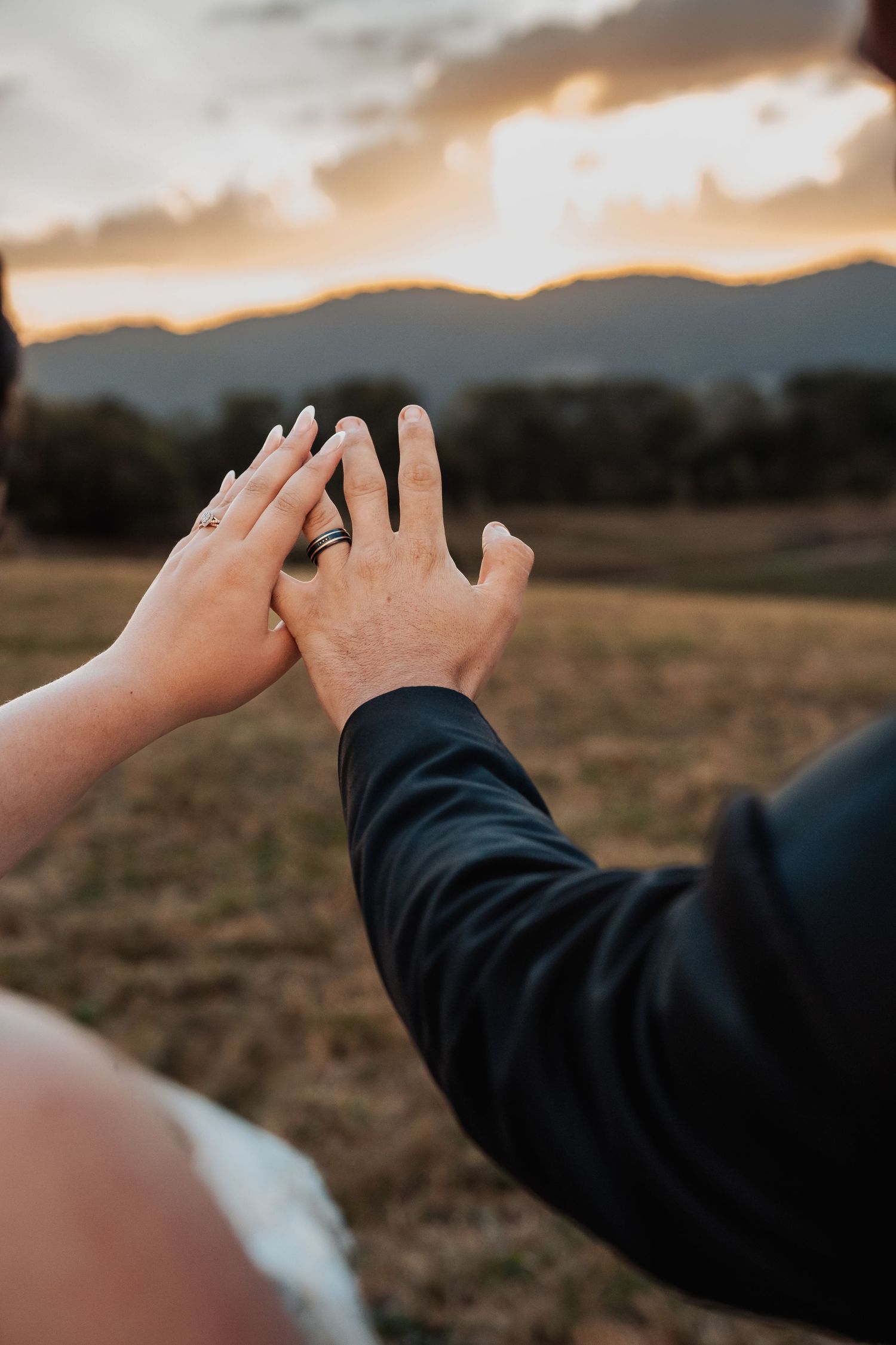 Bride and groom touching hands at sunset, showing wedding rings, with mountain landscape in background.