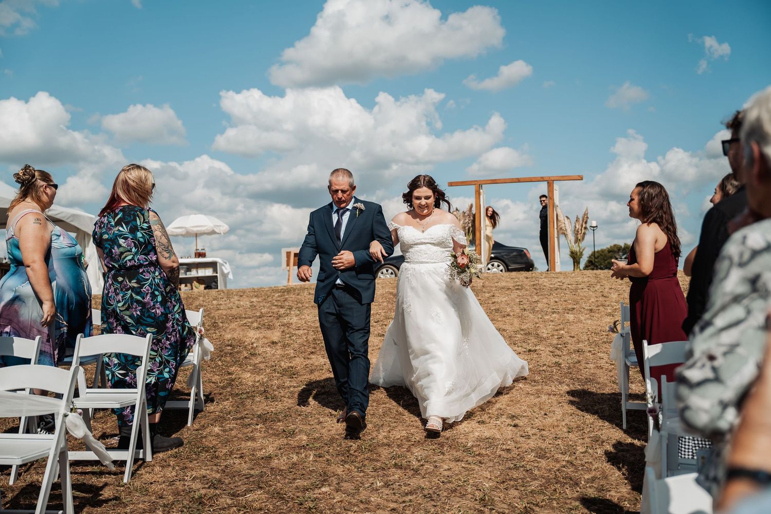 Bride in white gown walks down outdoor aisle with escort past seated guests under a bright blue sky.