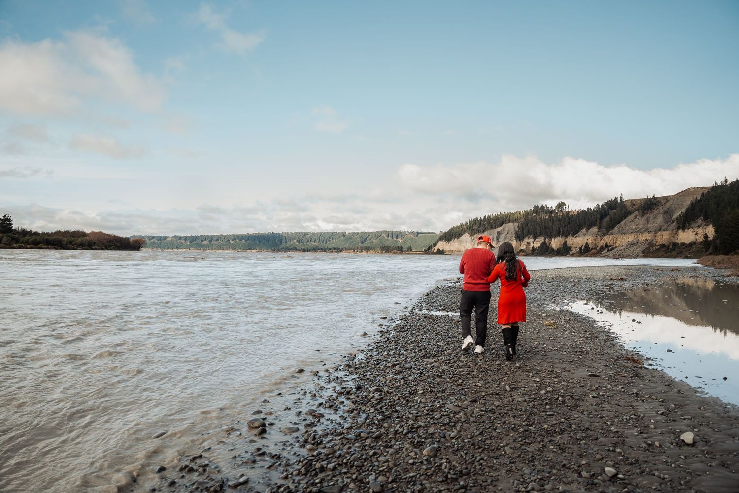 Couple in red outfits walking hand in hand along a rocky riverbank with forested hills in the background.