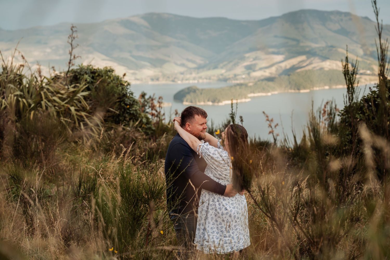 Couple embracing in tall grass with scenic lake and mountain backdrop during golden hour outdoor photoshoot.