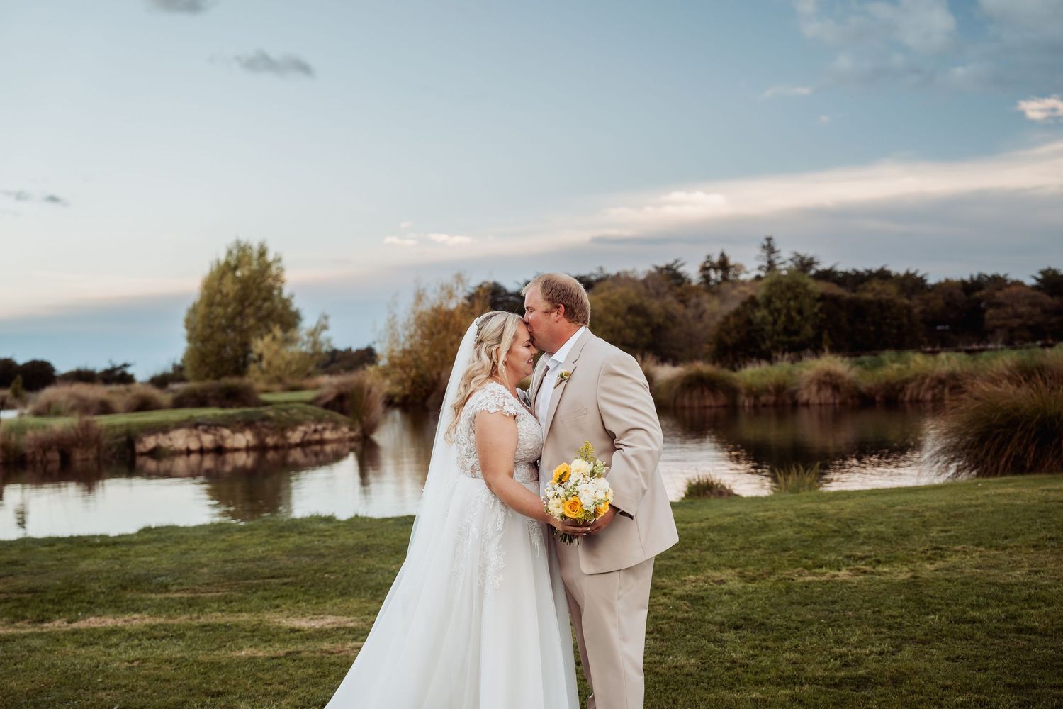 Bride and groom sharing a kiss in front of a scenic pond on their wedding day, bride holding yellow bouquet.