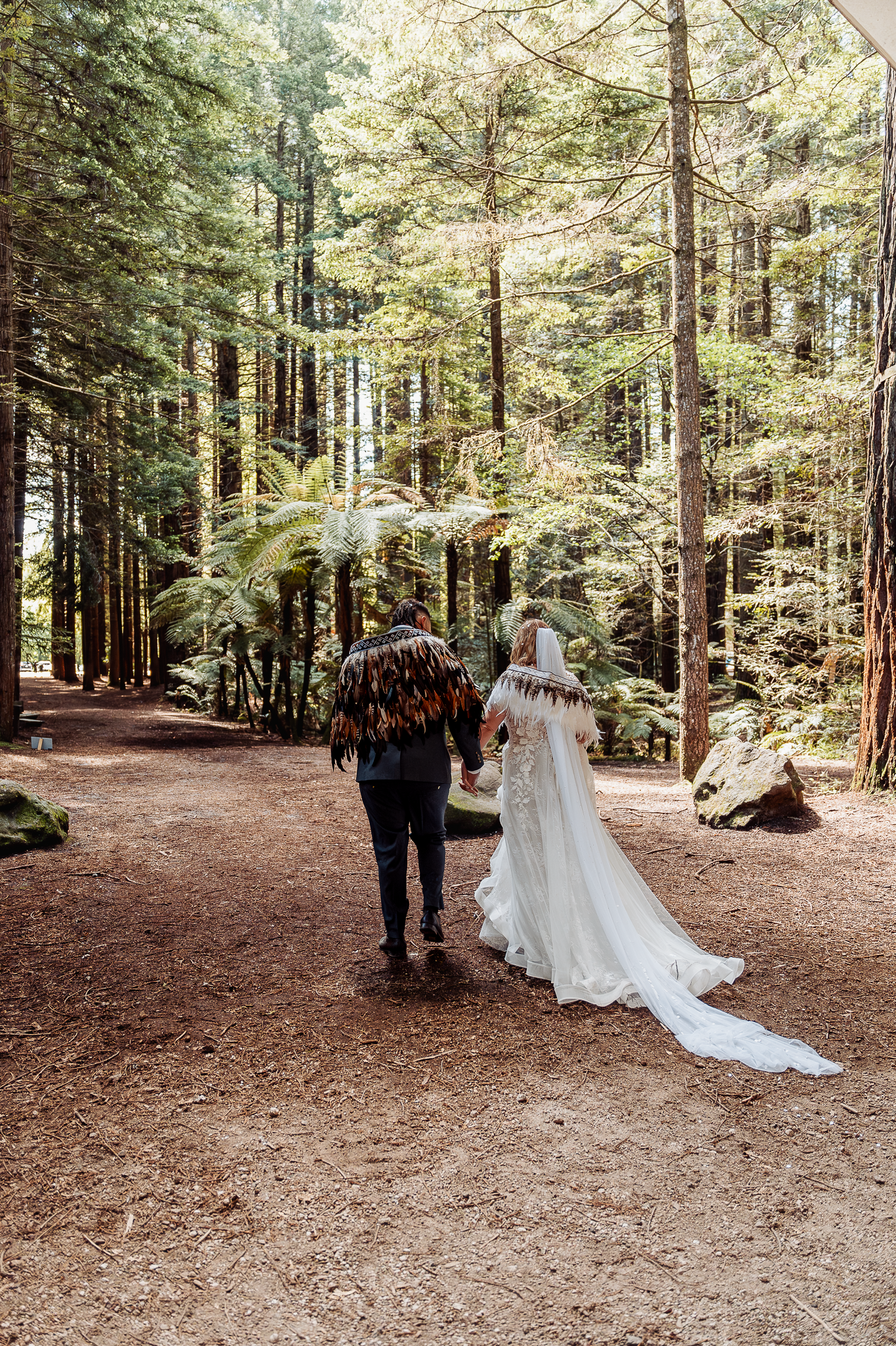 Wedding couple walking through forest path with tall trees.