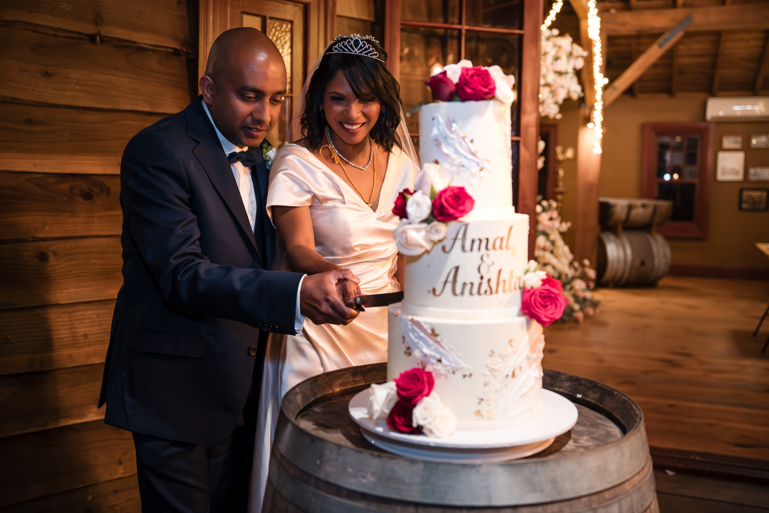 A couple cuts into their elegant white and pink wedding cake displayed on a rustic wooden barrel.