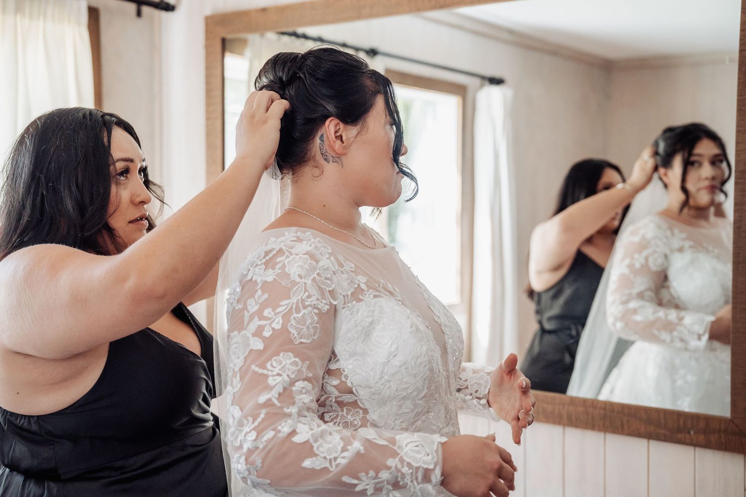 Wedding preparation scene with bride getting ready in elegant lace dress while helper assists with final touches.
