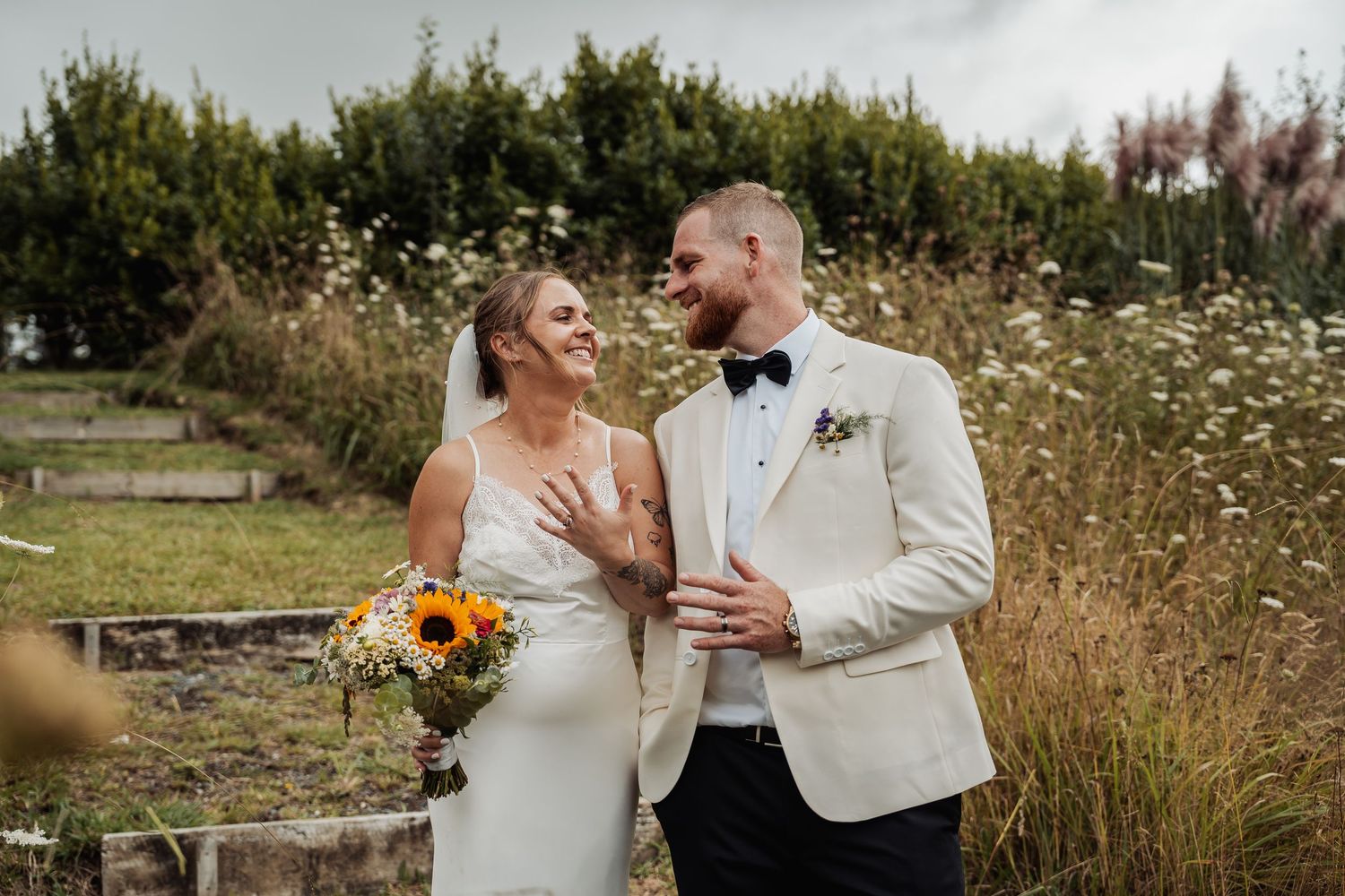 Bride and groom smiling at each other outdoors, she holds sunflowers, he wears a white tuxedo jacket with boutonniere.
