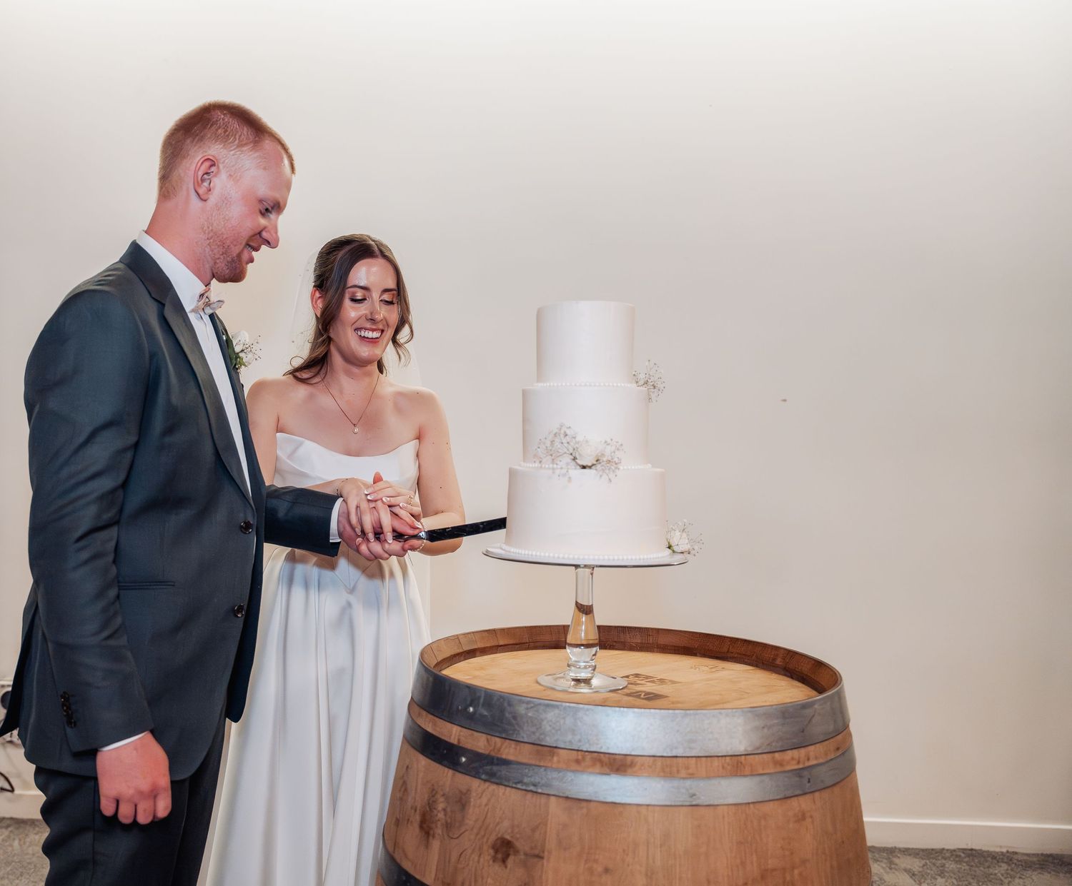 Smiling bride and groom cutting a three-tier white wedding cake displayed on a wooden wine barrel.