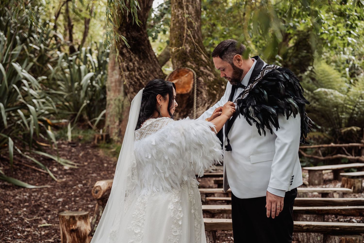 Bride adjusts grooms feathered jacket lapel during intimate outdoor forest wedding ceremony.