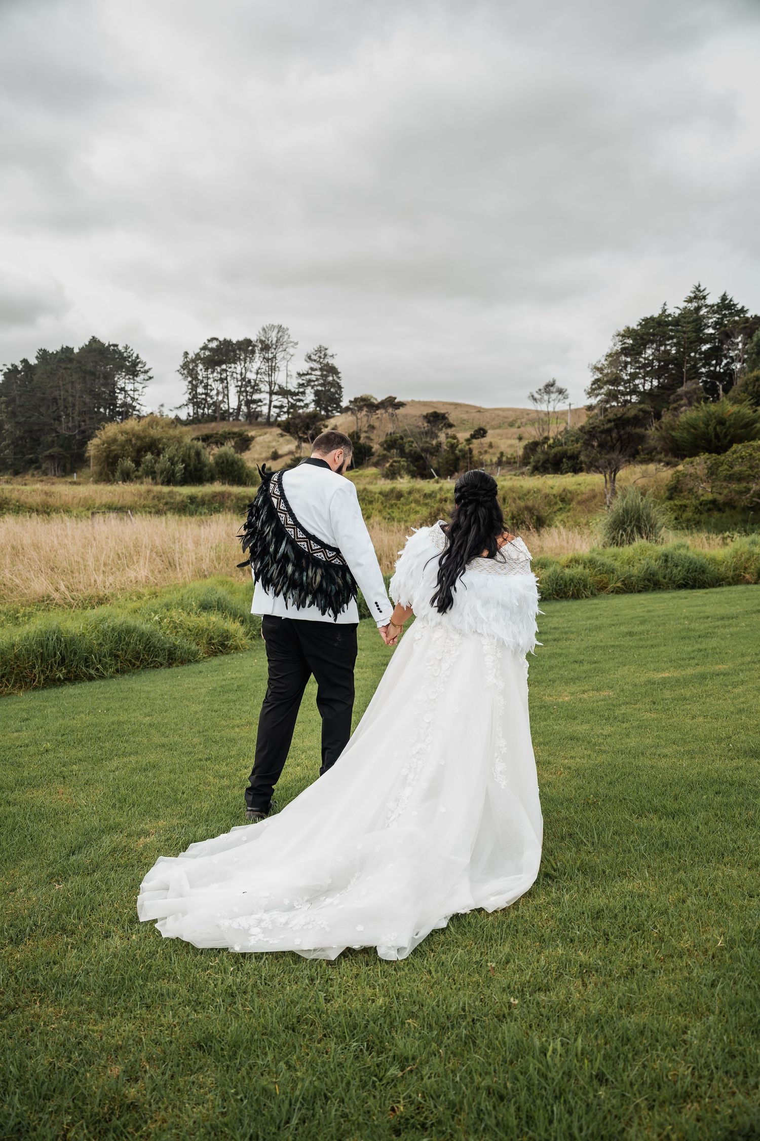 Bride and groom in feather-adorned wedding attire walk hand in hand across a lush green rural landscape.