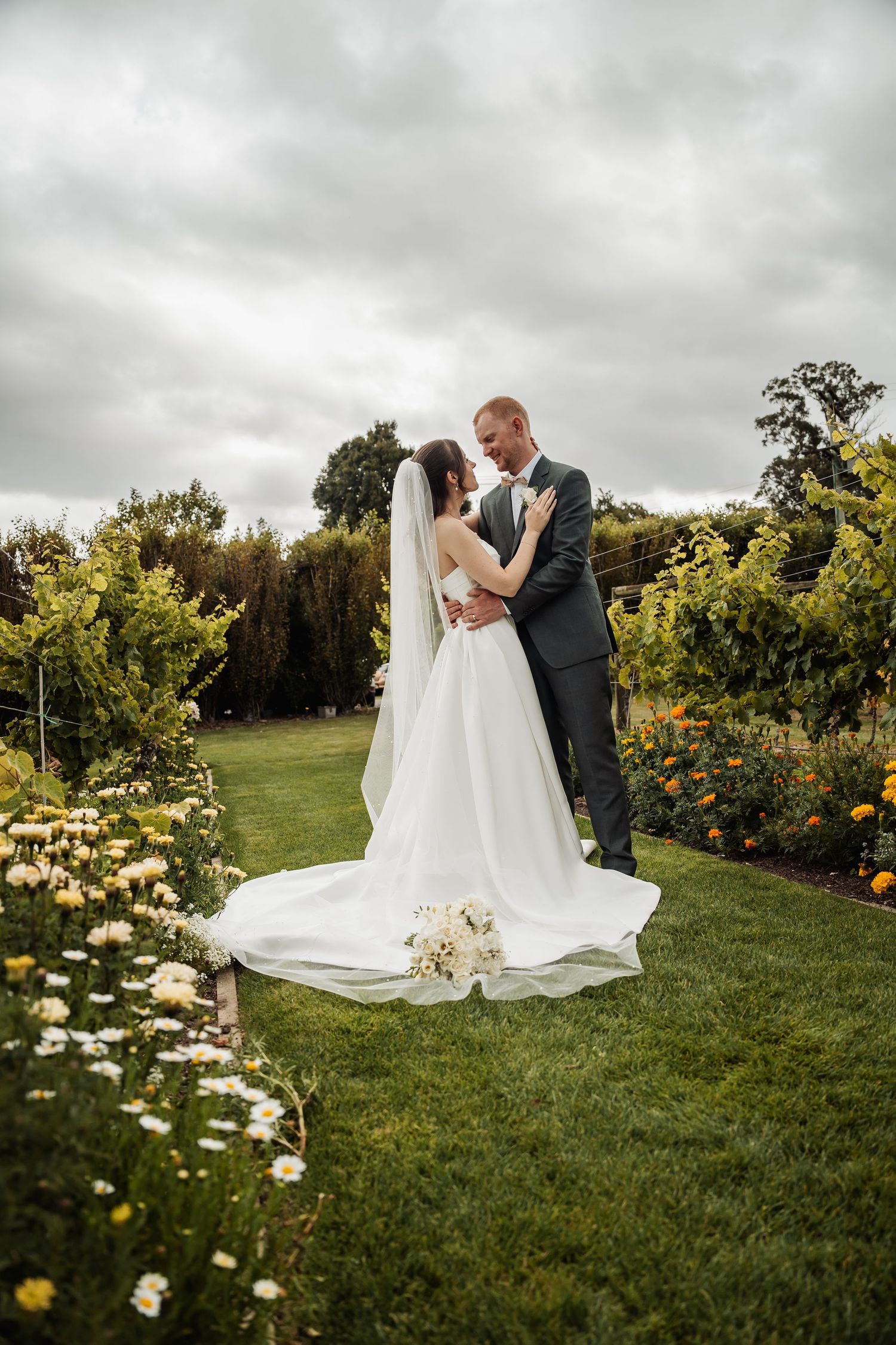 Bride and groom embrace in lush garden surrounded by colorful flowers and grapevines under cloudy sky.