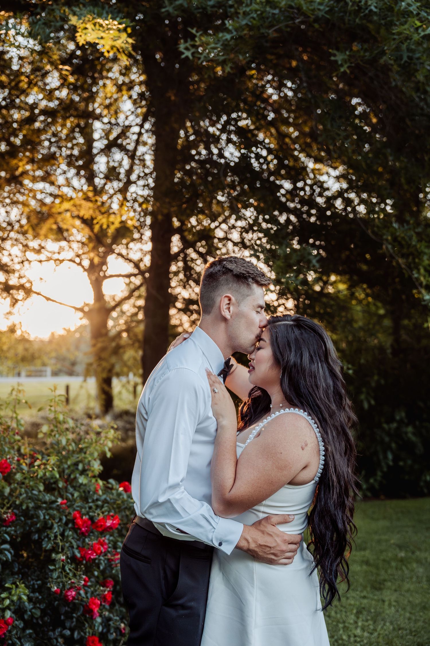 Bride and groom sharing a romantic sunset kiss near red roses in a garden setting.