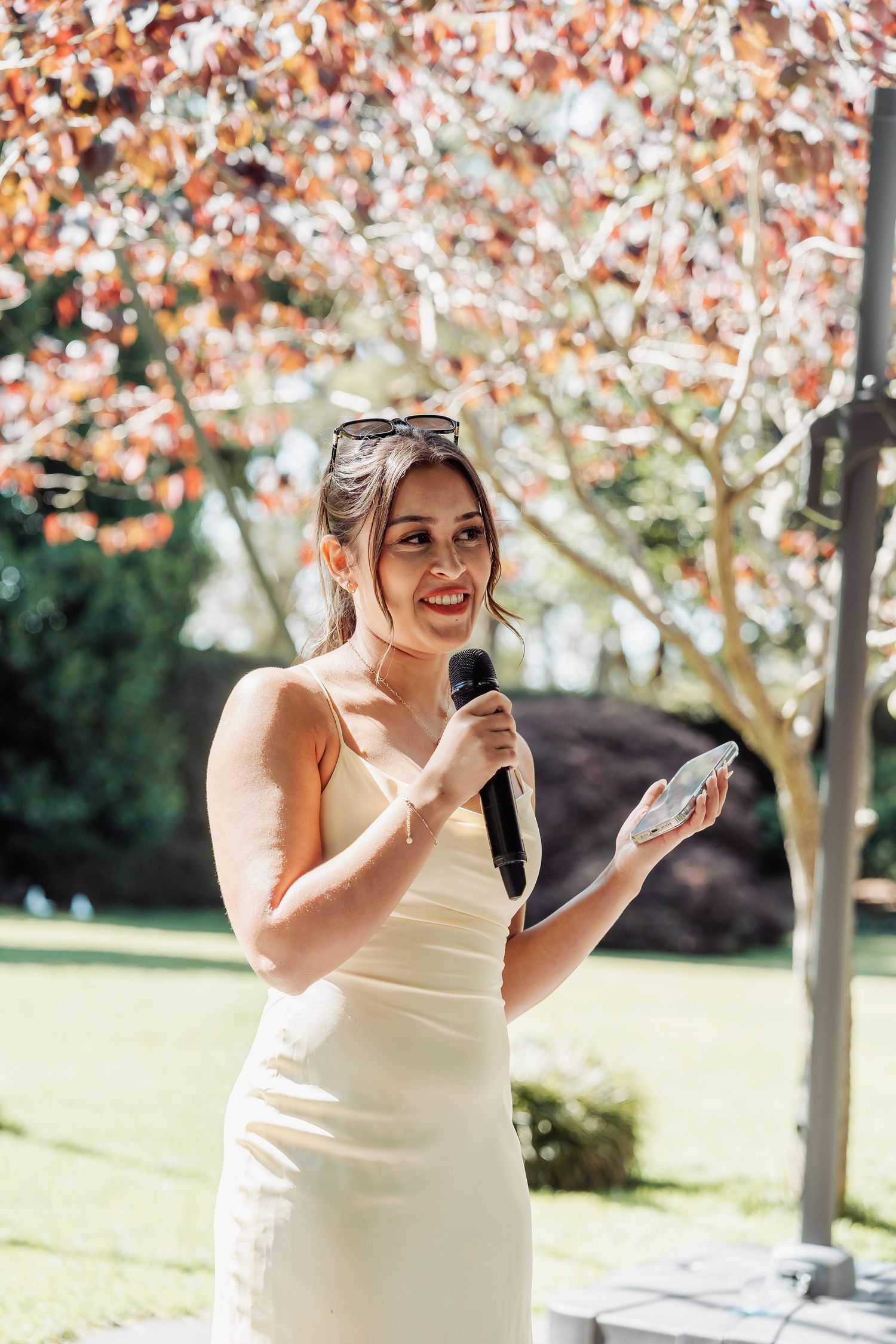 Smiling bridesmaid in white dress gives speech holding microphone and phone beneath colorful autumn tree outdoors.