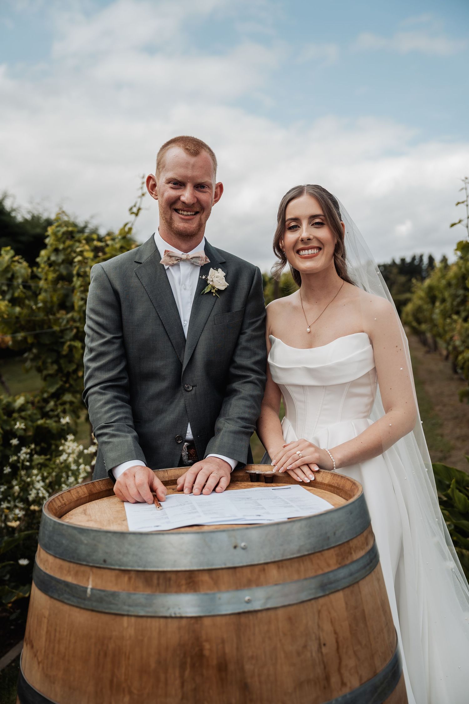 Smiling newlyweds sign their marriage certificate on a rustic wine barrel in a lush vineyard setting.