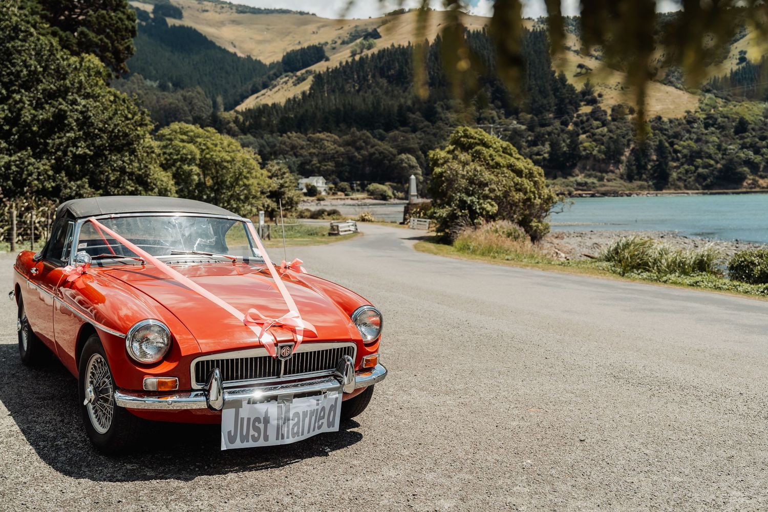 Red MG convertible wedding car with Just Married sign parked on scenic New Zealand road near lake and mountains.