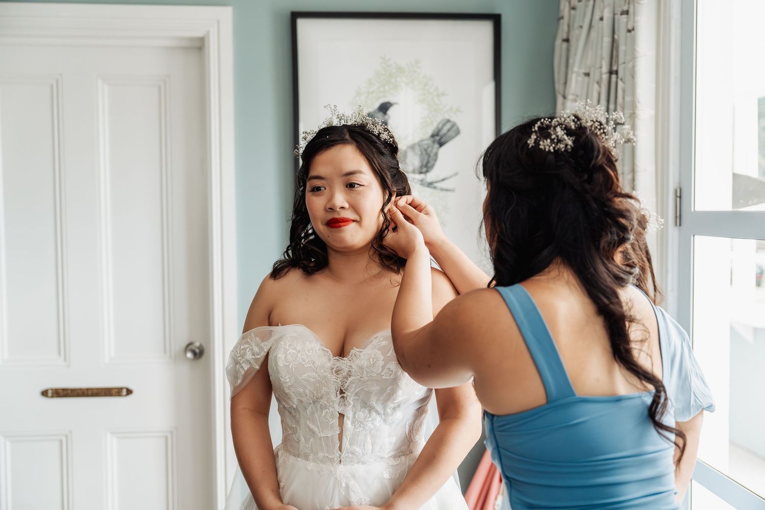 Bridesmaid in blue dress helping Asian bride in lace wedding gown with earrings in bright dressing room.