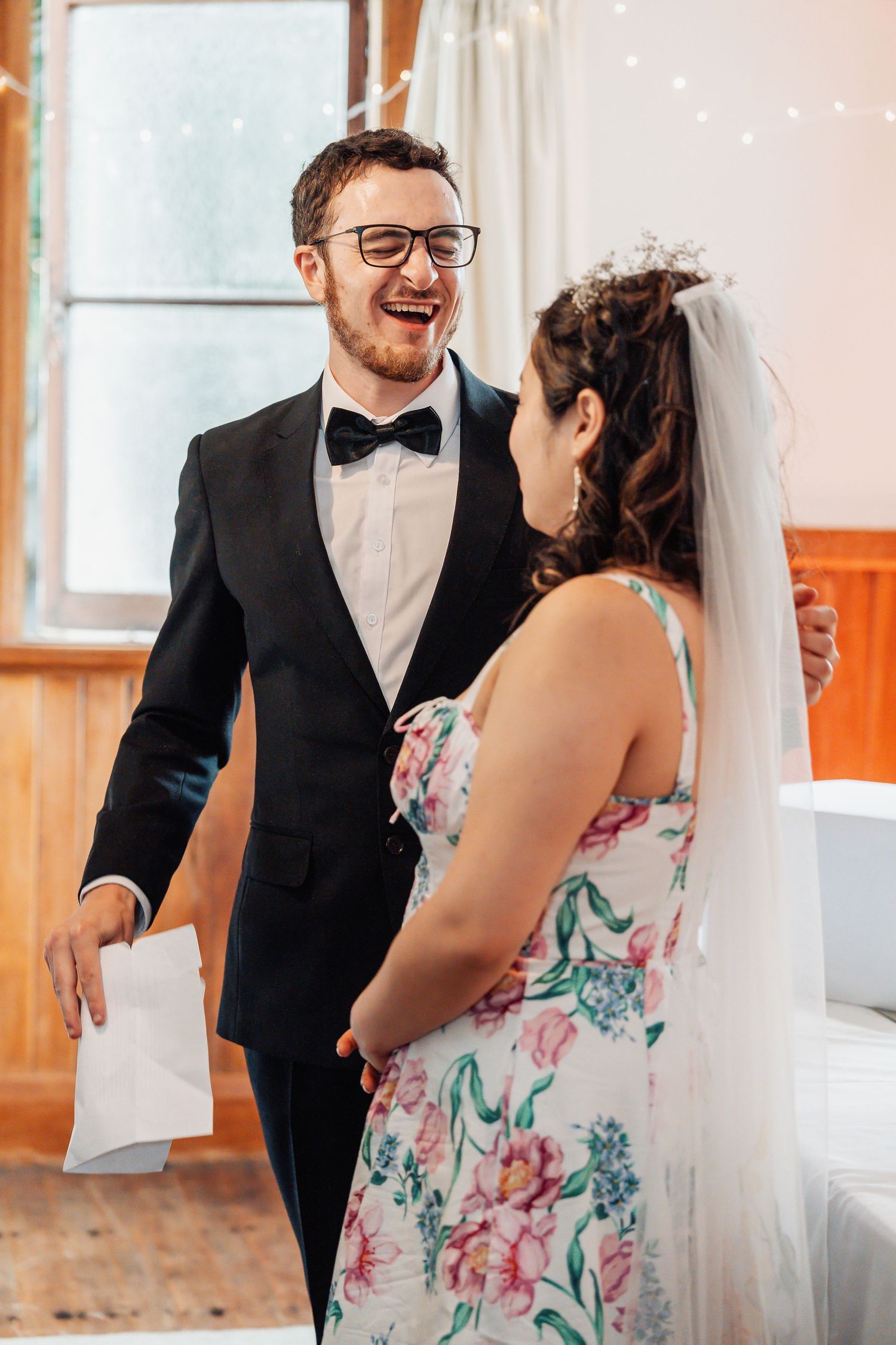 Laughing groom in tuxedo shares joyful moment with bride in floral dress at indoor wedding.