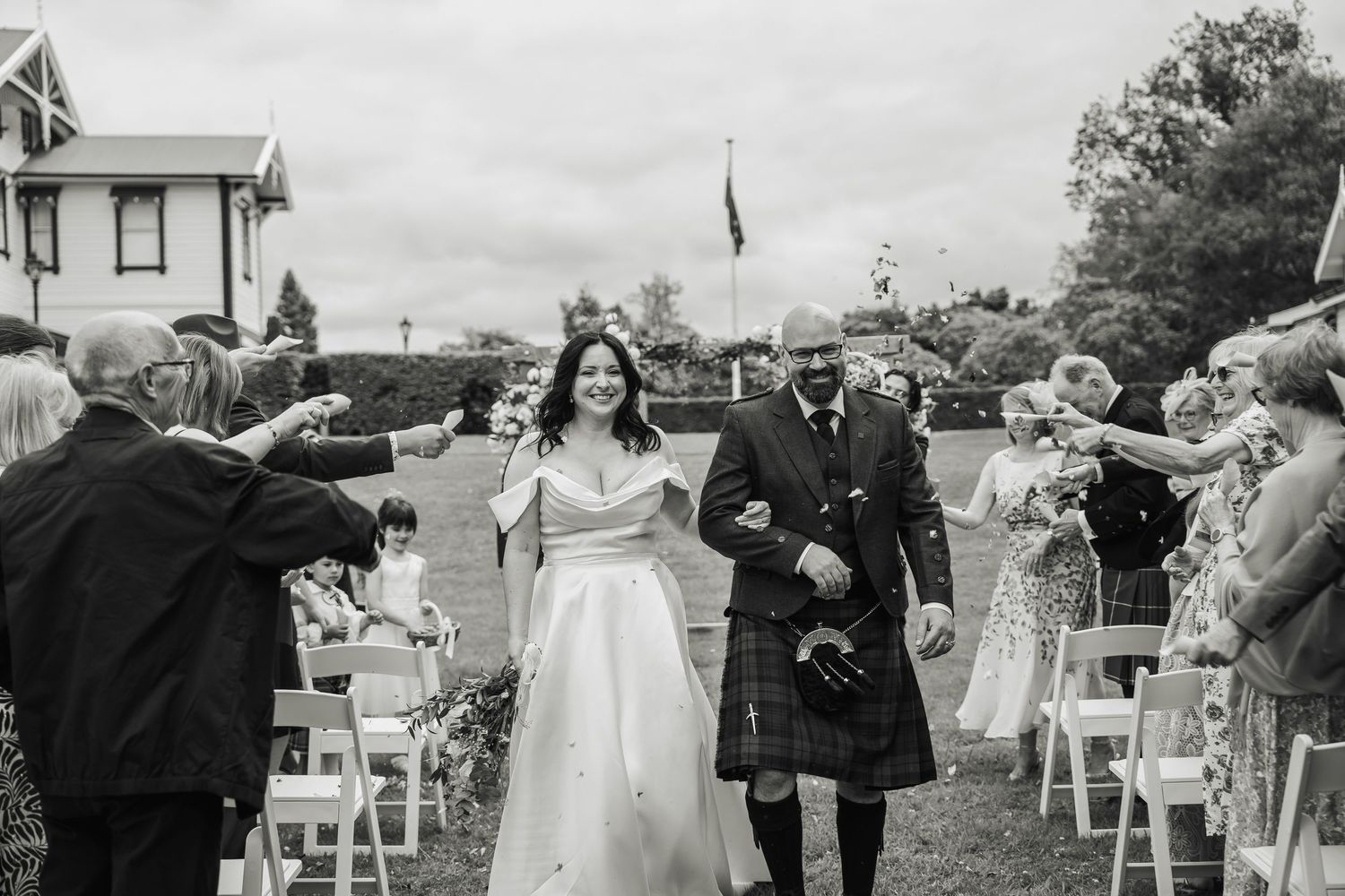Bride and kilted groom walk back down the aisle as guests celebrate in black and white.