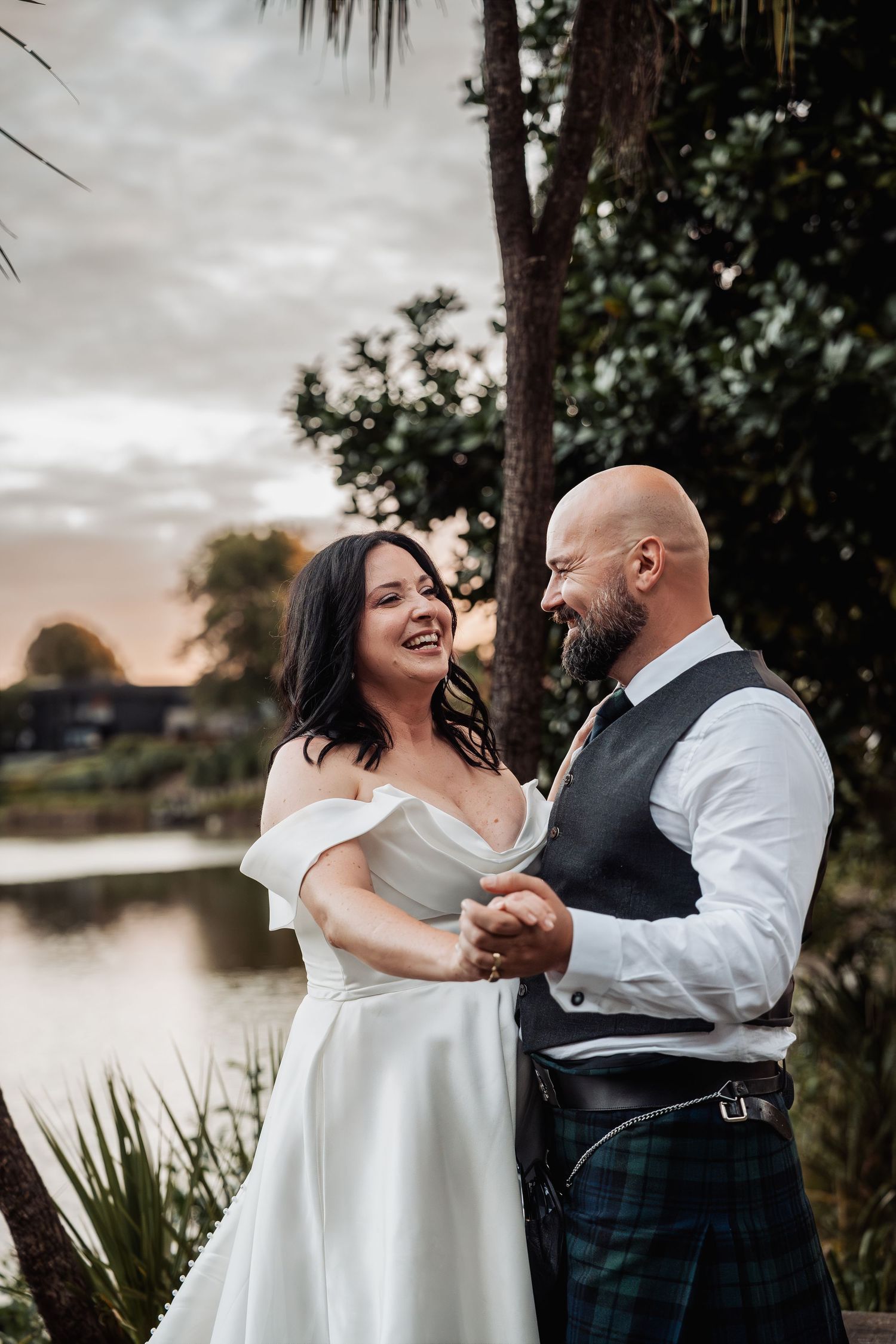 Laughing bride in white off-shoulder gown dances with bearded groom in tartan kilt by a lake at sunset.