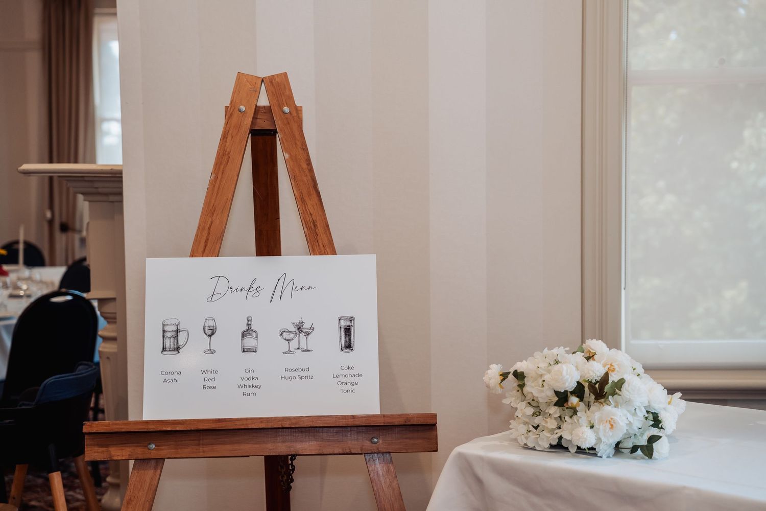 Wooden easel displaying a drinks menu sign at a wedding reception, with a white rose bouquet on a nearby table.